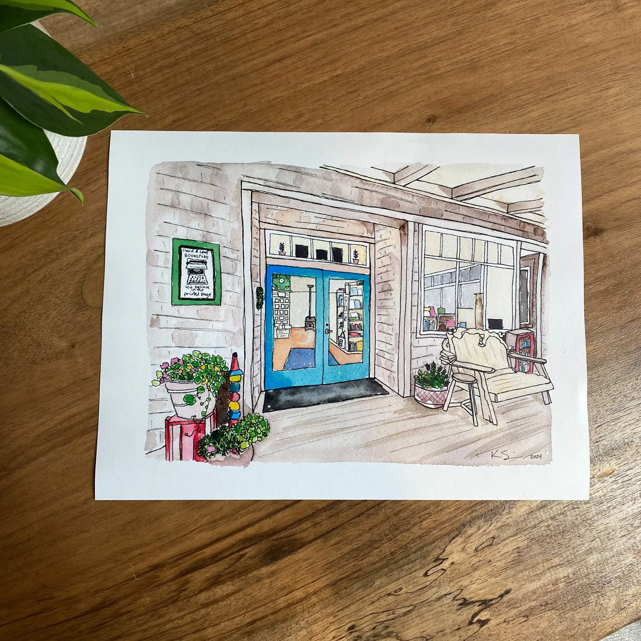 Cloud & Leaf Bookstore watercolor print on 8.5x11 premium paper, on a wooden desk, featuring soft tones and lively accents that bring this beloved coastal bookshop to life.