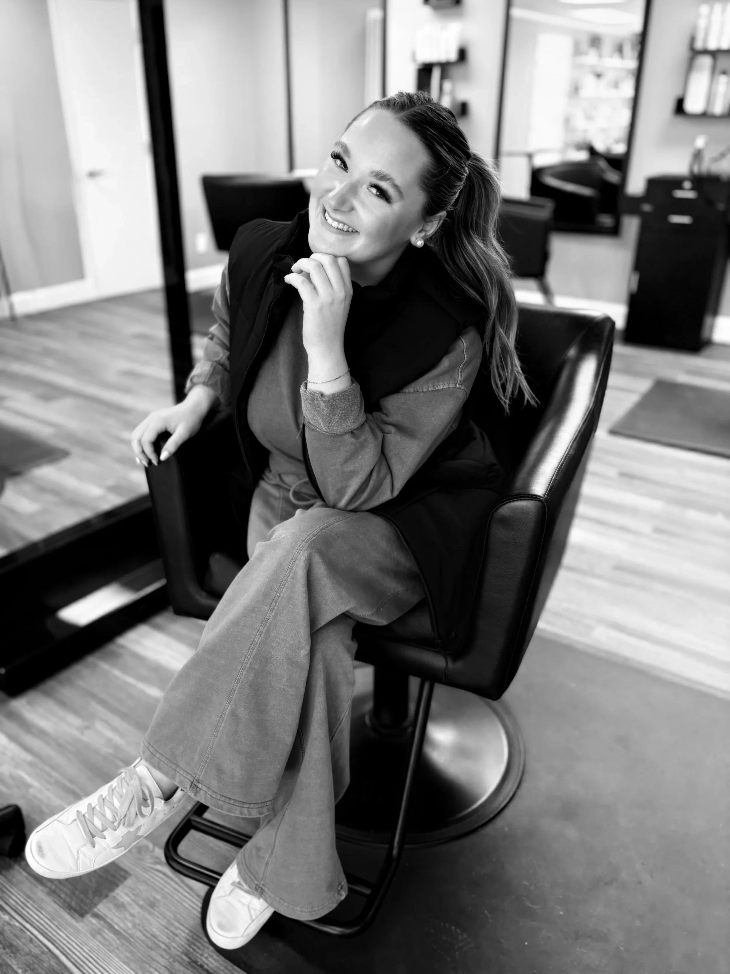 A smiling woman with long hair sitting in a salon chair in a hair salon, wearing casual clothes and sneakers, with hairdressing tools and salon decor visible in the background.