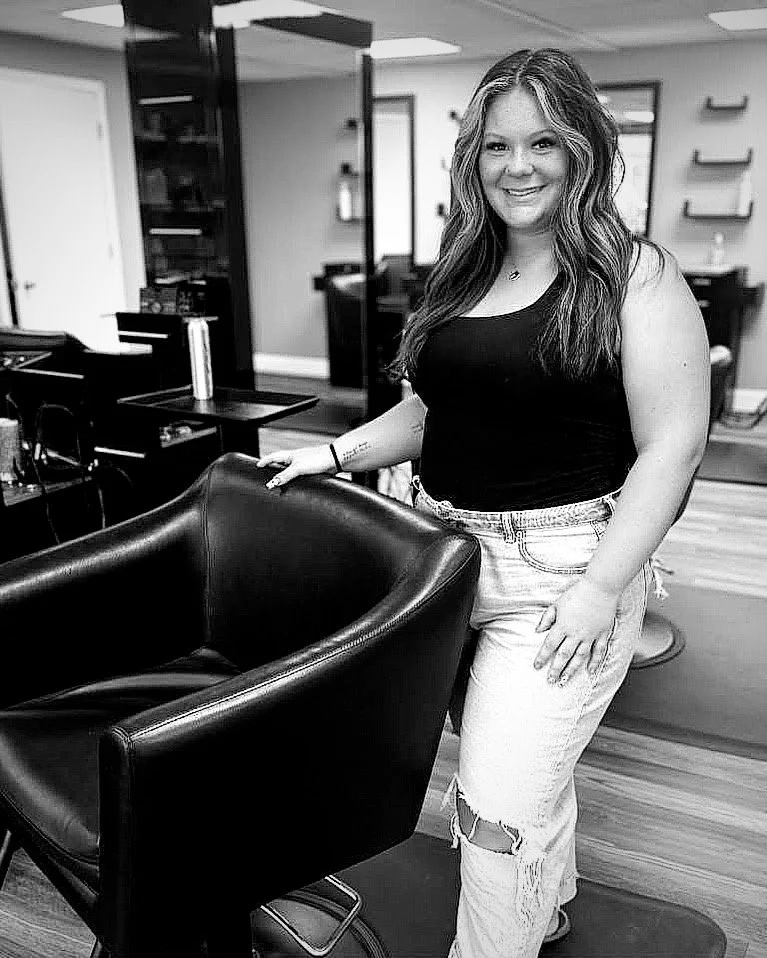 A young woman with long wavy hair smiling, standing in a hair salon, touching a black salon chair, wearing a black tank top and ripped jeans.