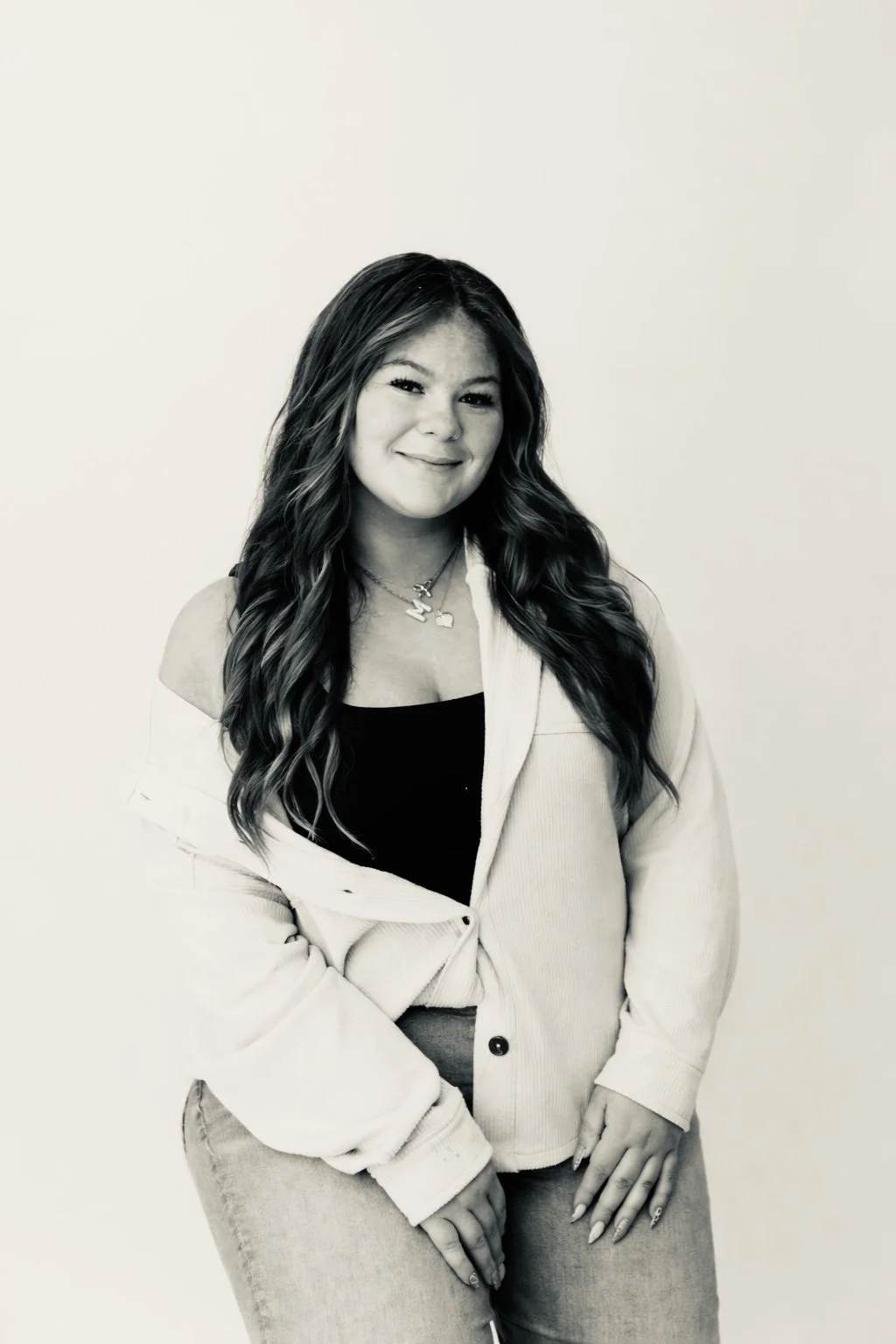 A young woman with long wavy hair, smiling, wearing a white jacket over a black top, with jewelry, standing against a plain light background.