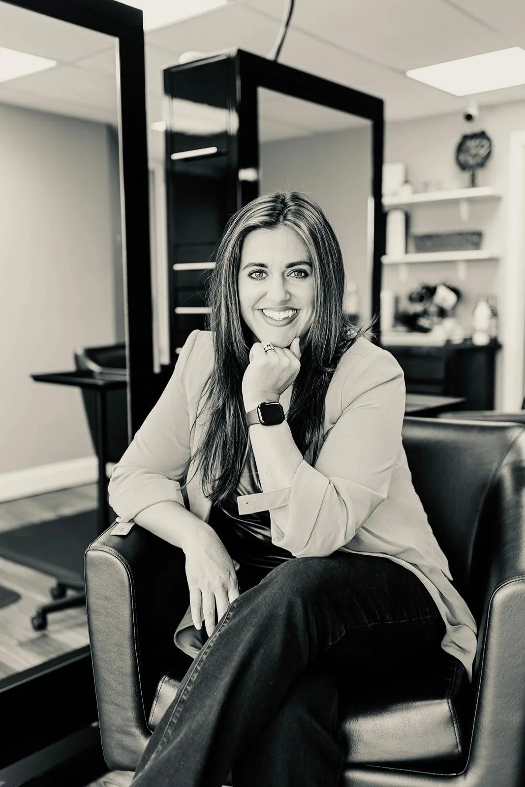 A woman with long hair sitting on a leather chair, smiling, with her chin resting on her hand, in an office setting.