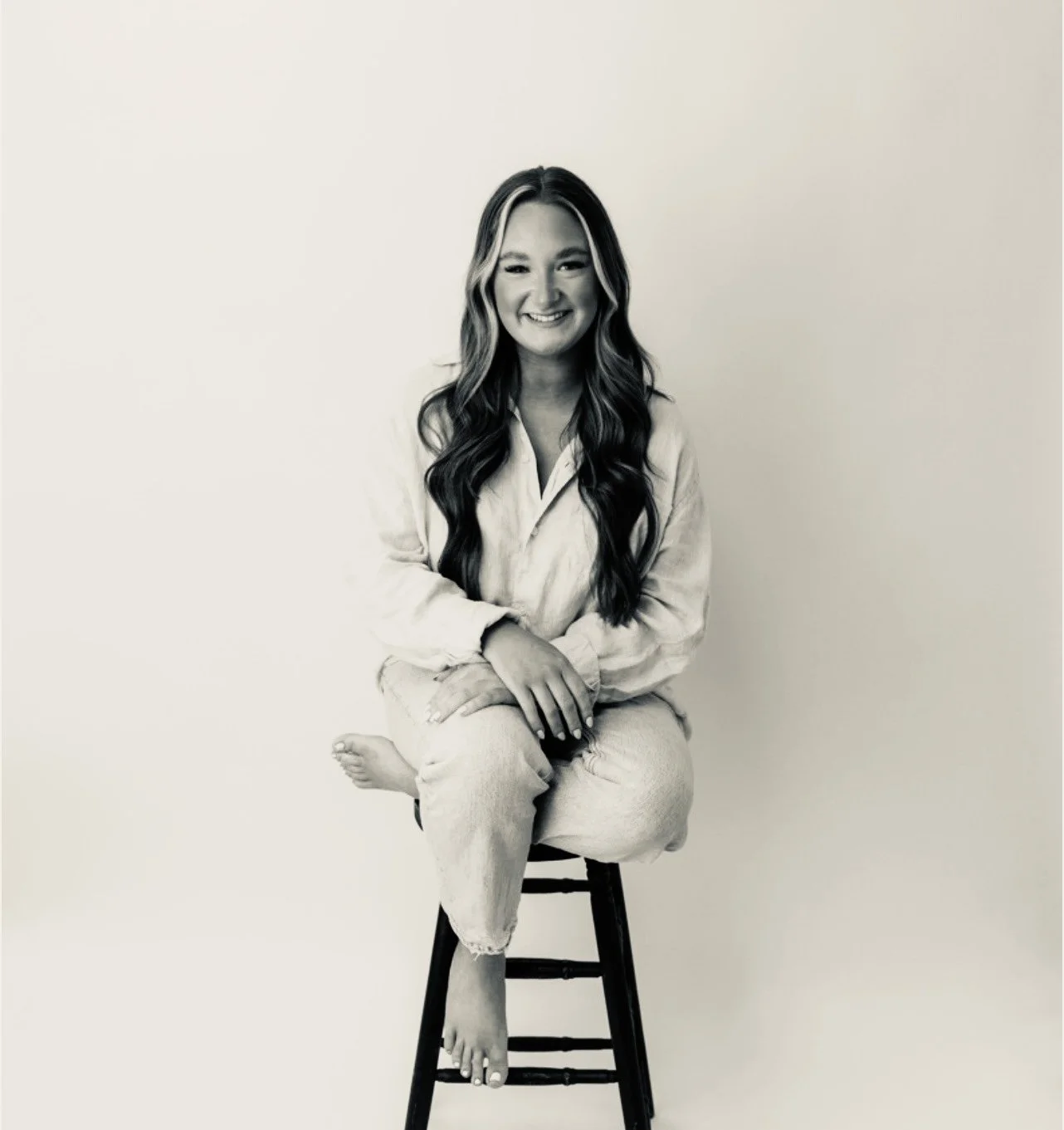 A young woman with long wavy hair sitting cross-legged on a bar stool, wearing a light-colored shirt and jeans, smiling at the camera, in a plain studio background.