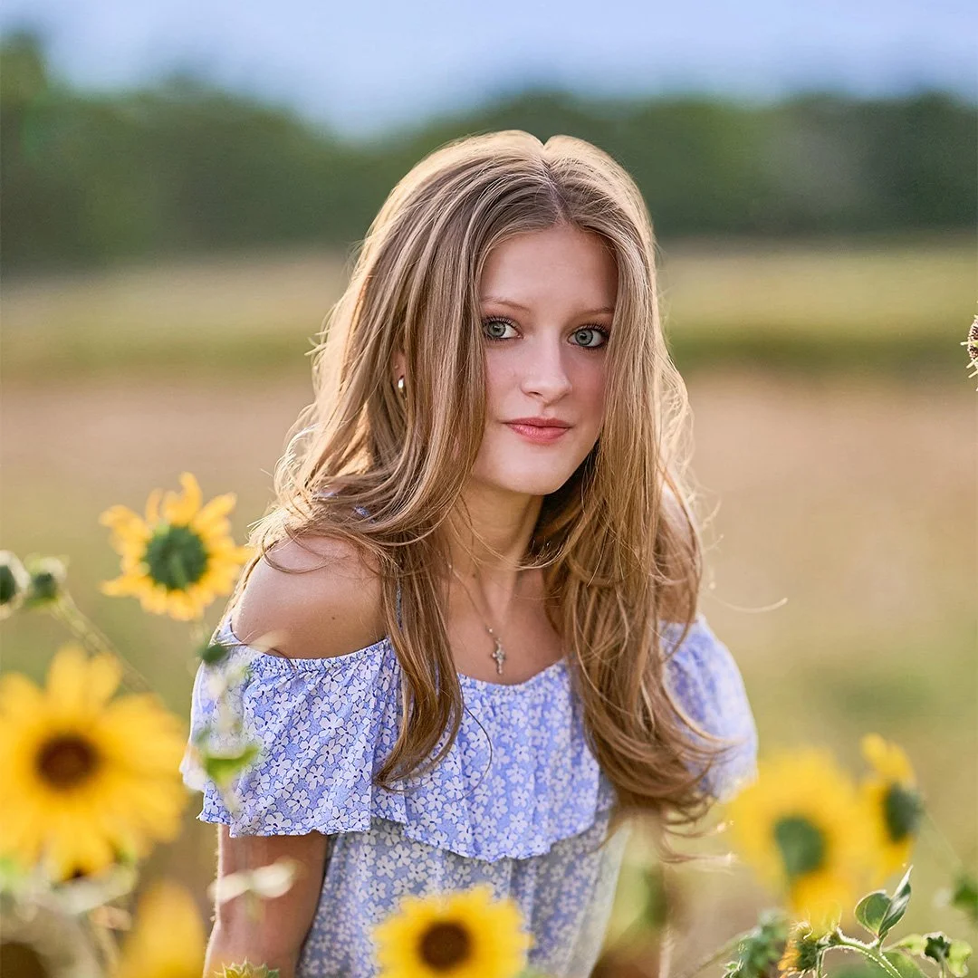 A young woman with long, wavy, light brown hair wearing a blue and white off-shoulder dress, standing outdoors among sunflowers in a field.