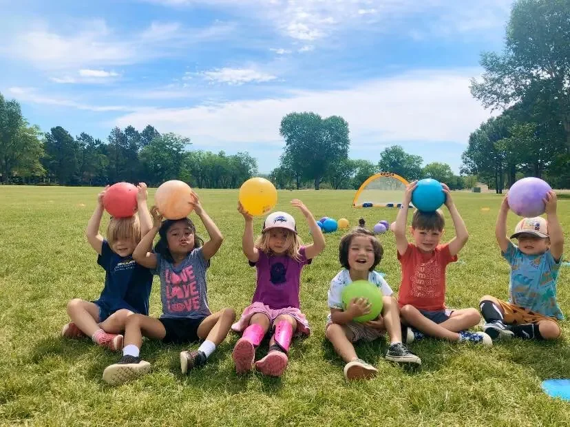 Six children sitting on grass in a park, holding colorful balls above their heads, with trees and blue sky in the background.