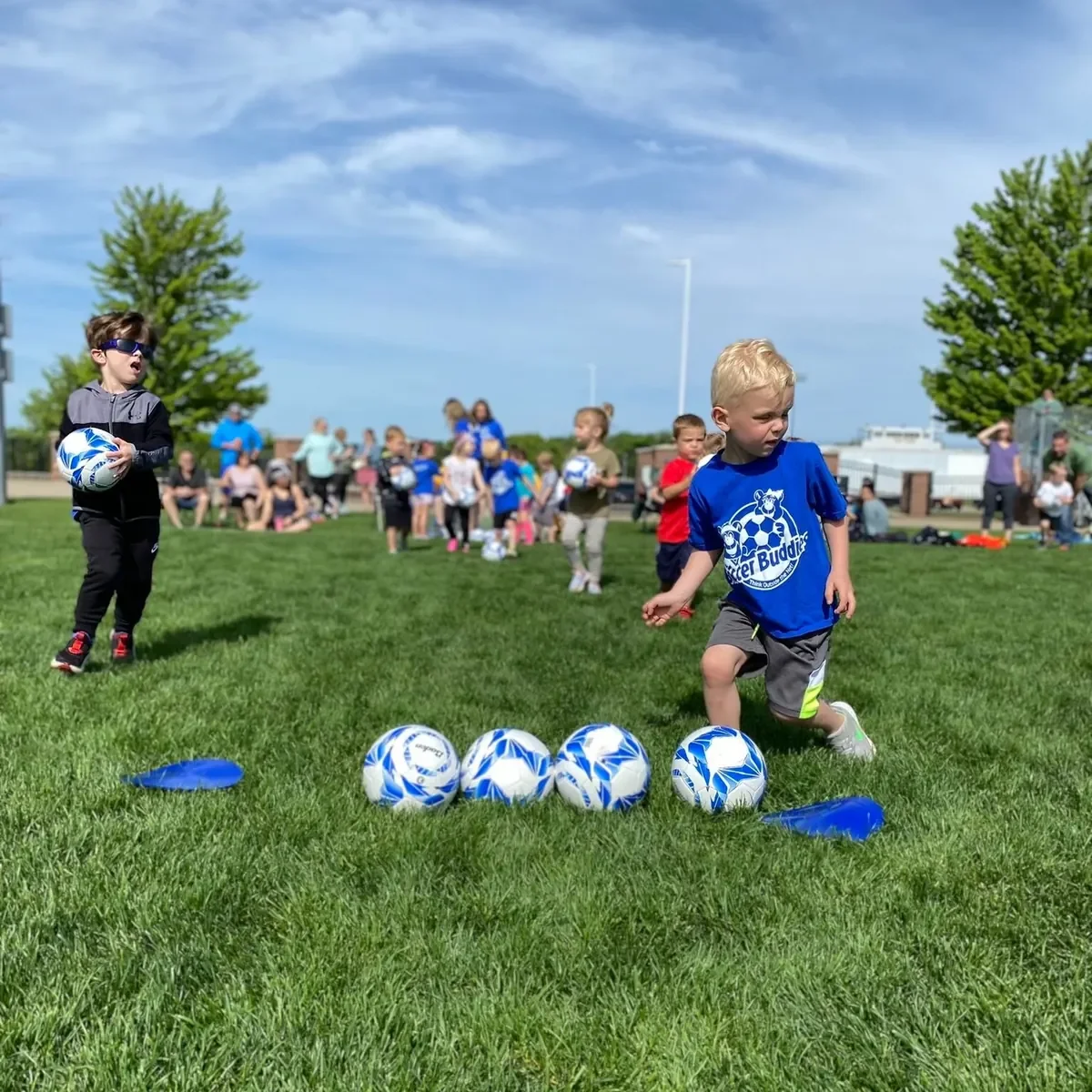 Group of children on a grassy field participating in a soccer practice or event, with kids in the foreground preparing for activity and others in the background, some sitting and others standing.