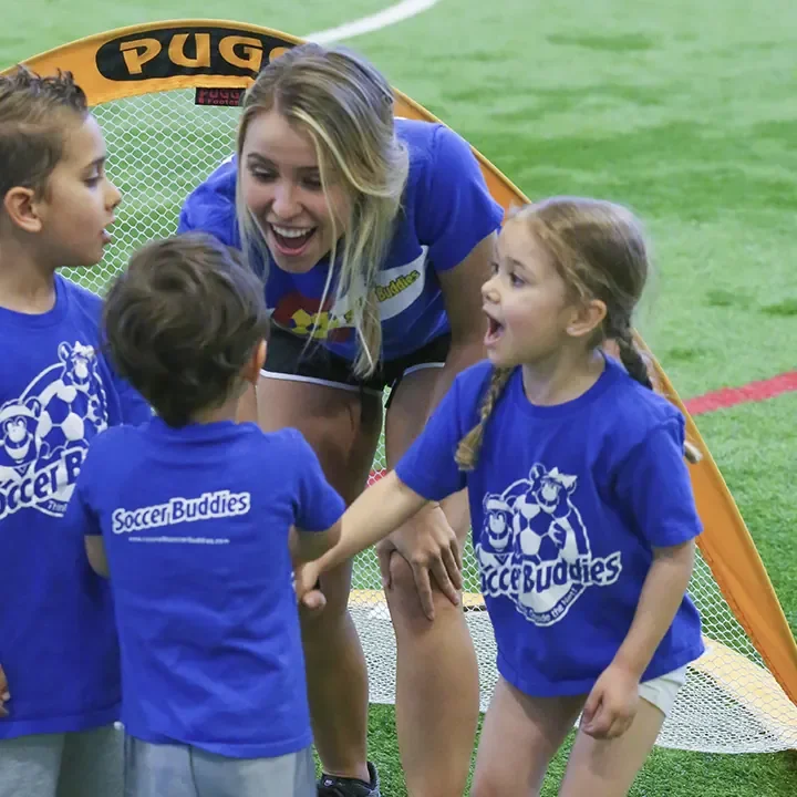 A female soccer coach talking to a group of young children on an indoor soccer field, all wearing blue Soccer Buddies shirts.