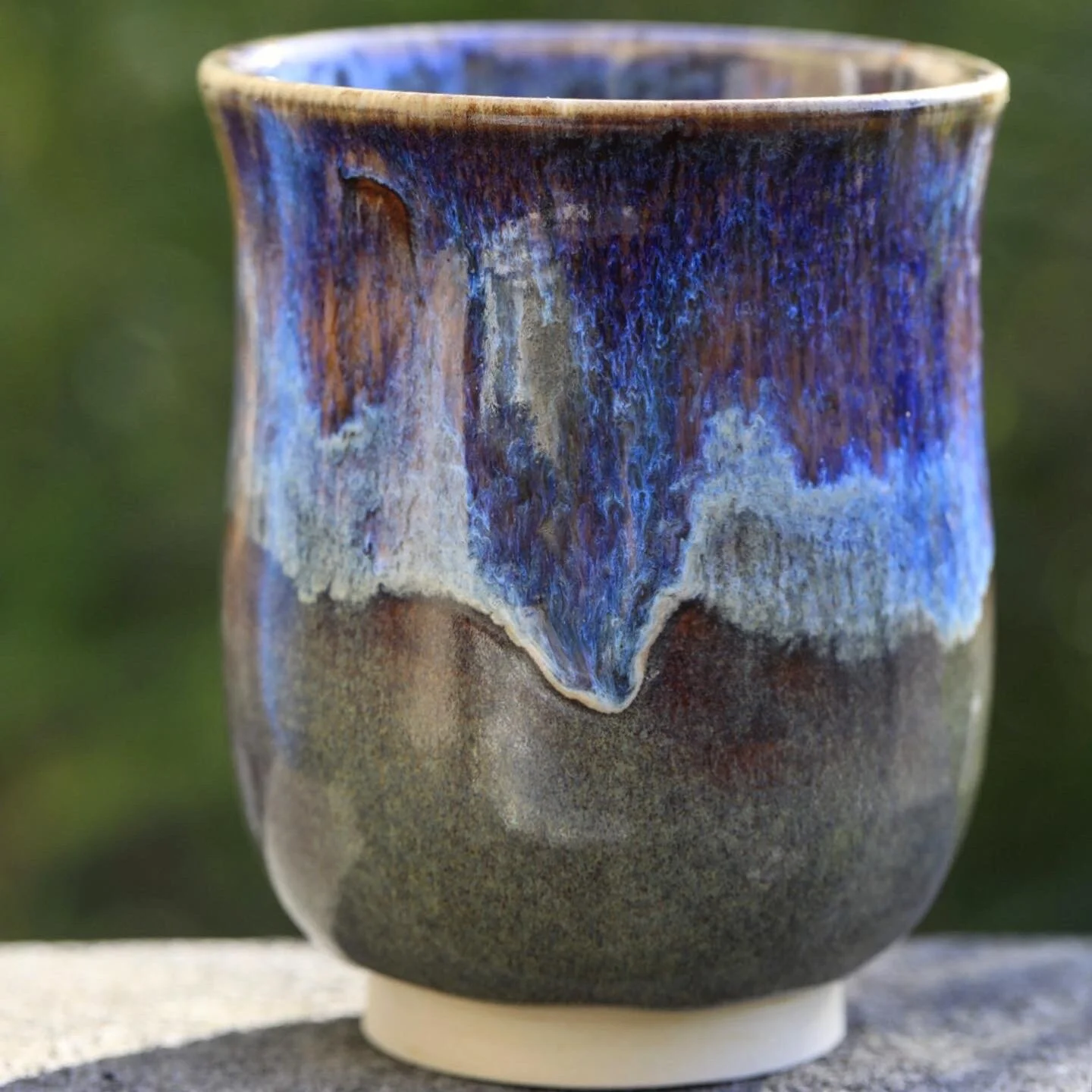 Close-up of a ceramic cup with a textured glaze featuring blue, purple, and gray colors, set outdoors on a flat surface.