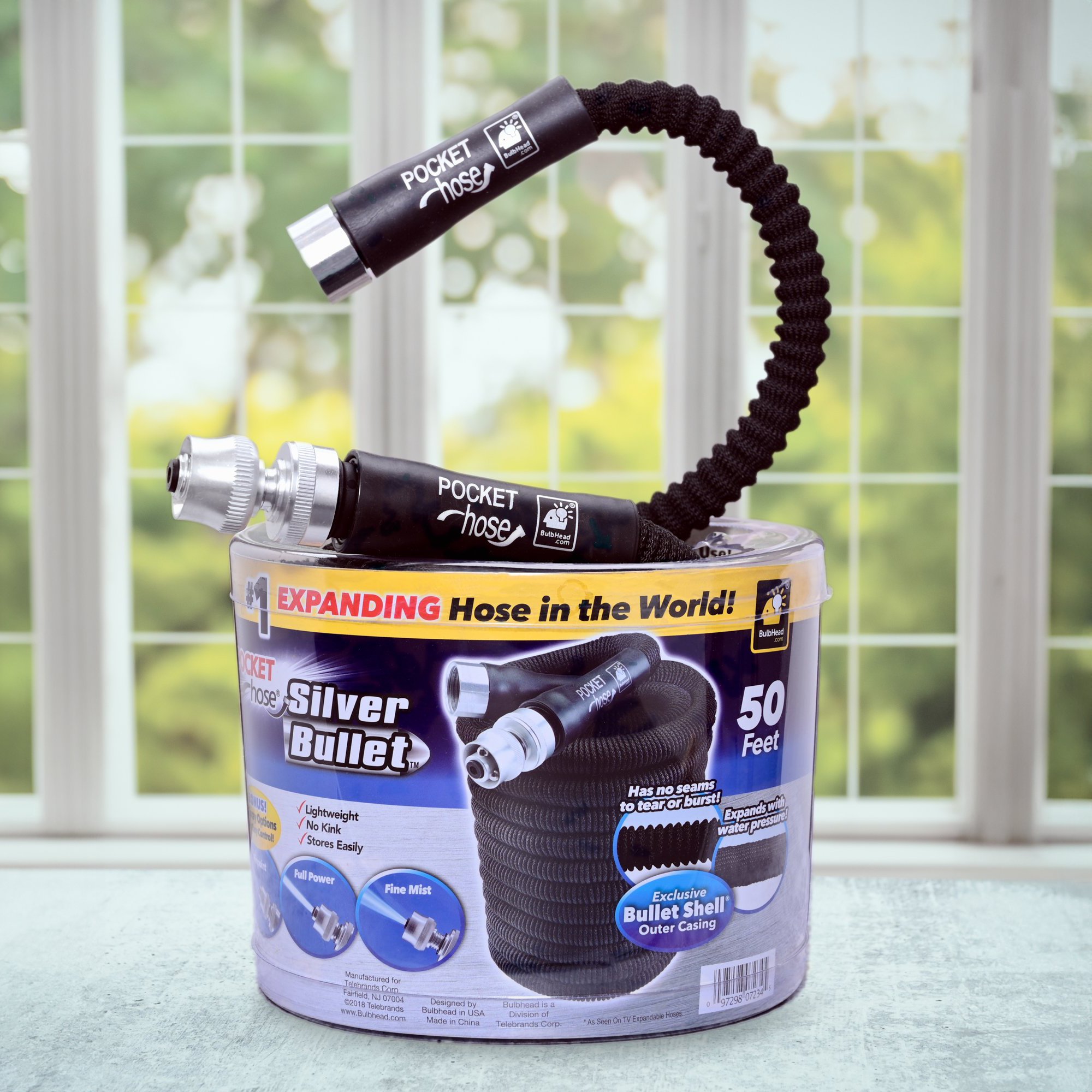  Flexible garden hose in the packaging on a kitchen countertop and windows with green trees outside. The hose is extending out of the plastic packaging showing off the included water nozzle  