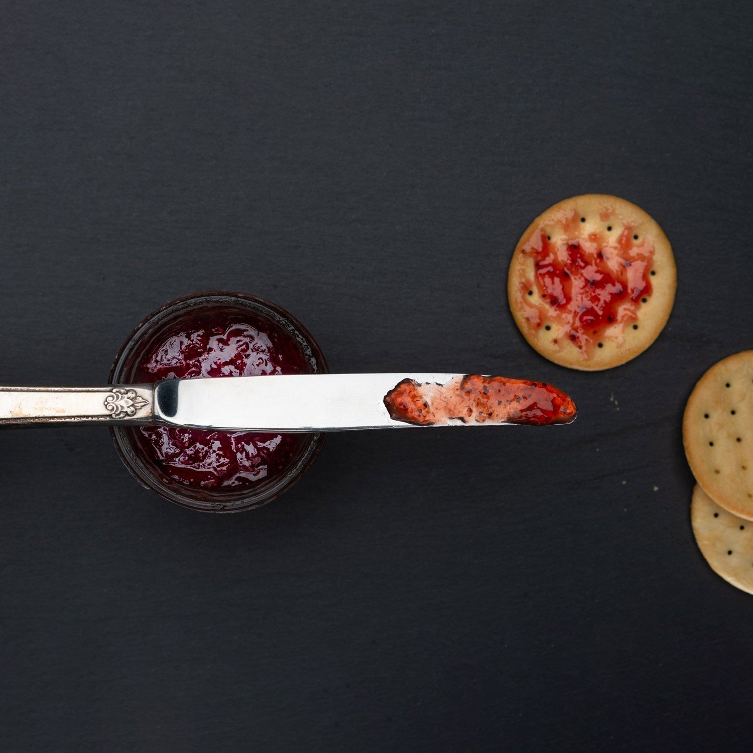  food photography of jelly on a knife blade sitting on a jelly jar with crackers on slate. 