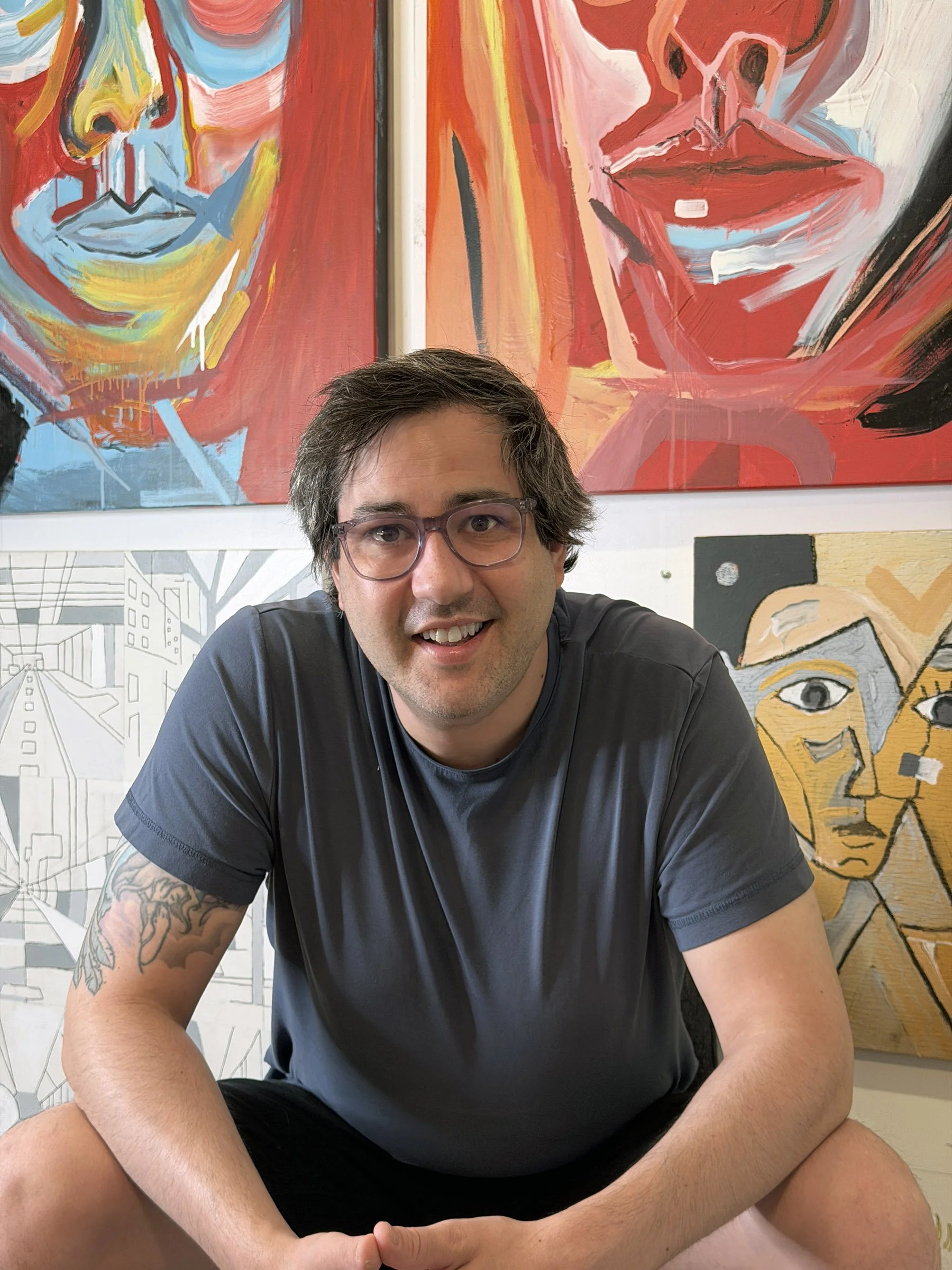 The artist sits with a happy sort of smile infront of his paintings. They are very colourful paintings and the artist is wearing violet glasses and a dark grey shirt.