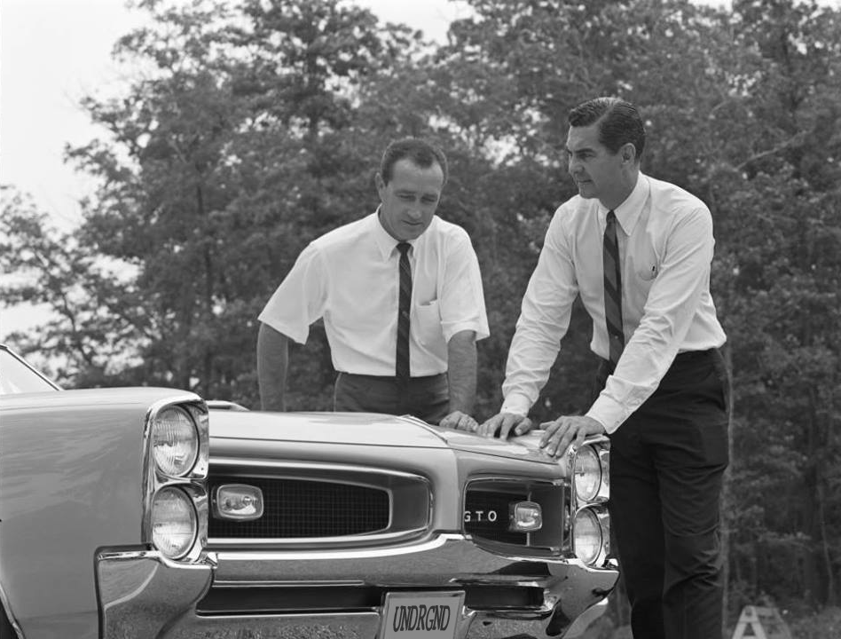 Two men in white shirts and ties examining a classic car outdoors, with trees in the background.
