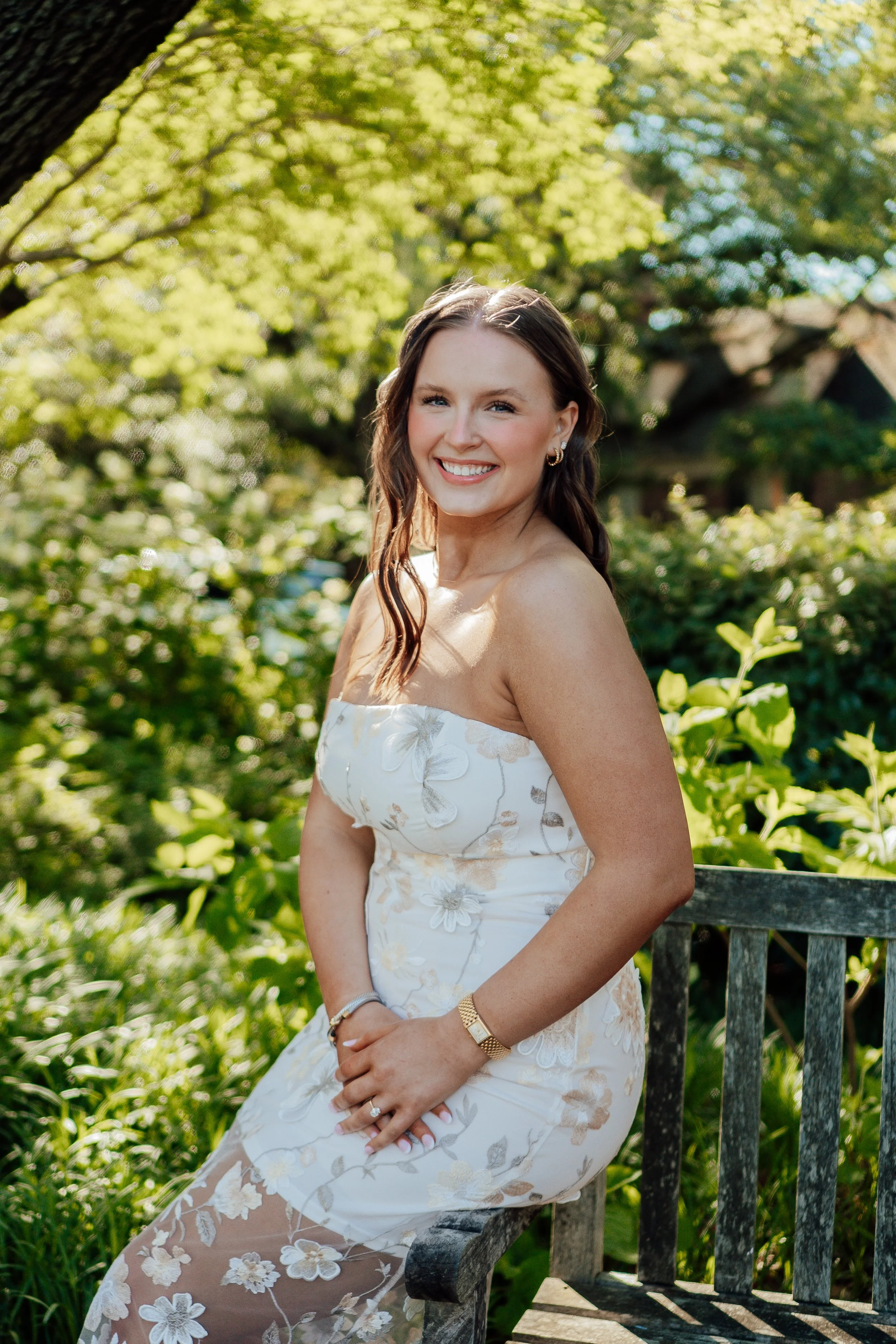 A woman with long brown hair smiling outdoors, wearing a white floral dress with strapless top, sitting on a wooden bench surrounded by green foliage and trees.