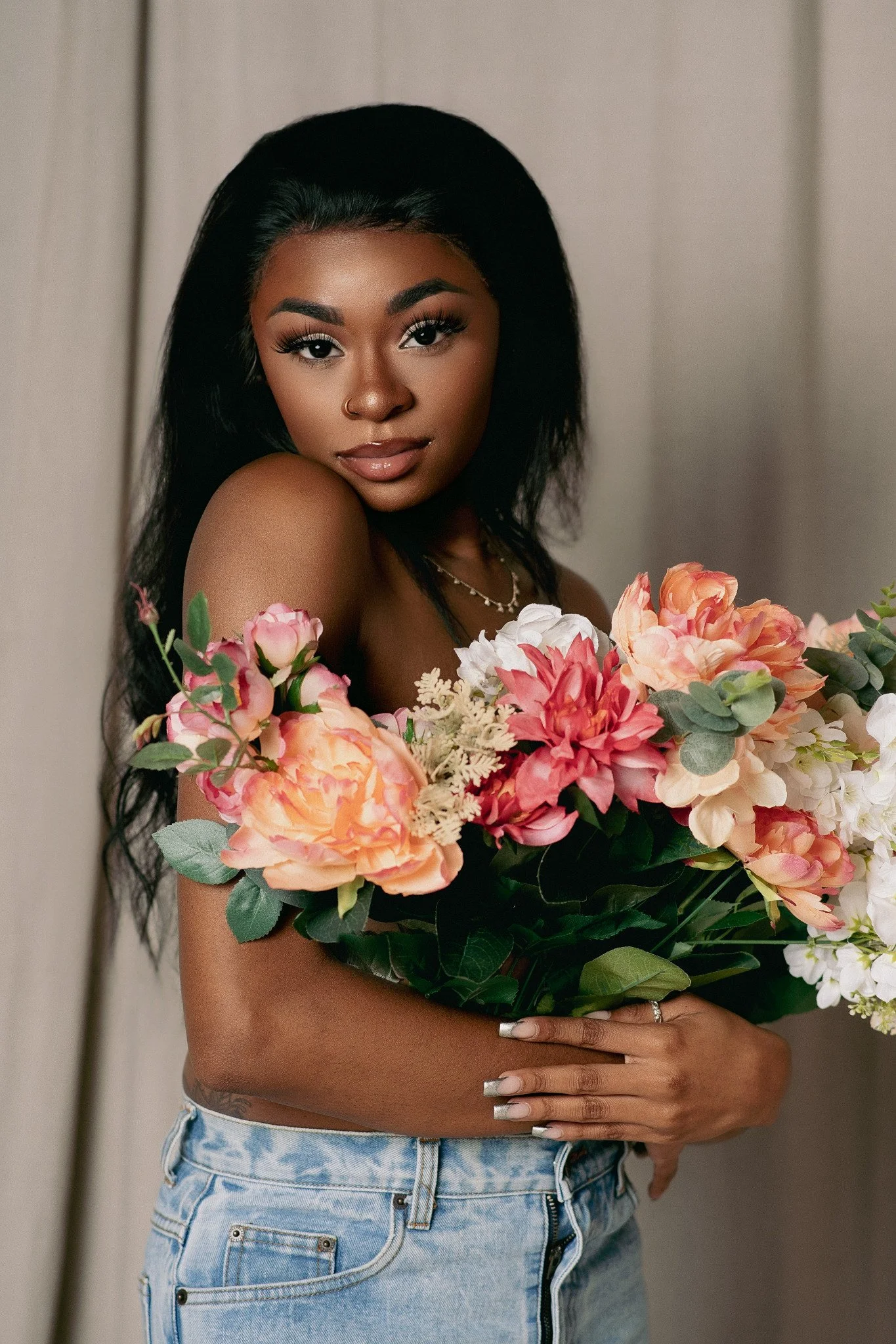 A woman with medium brown skin and long black hair holding a large bouquet of pink, peach, and white flowers, wearing a necklace, ring, light-colored denim shorts, standing against a plain light background.