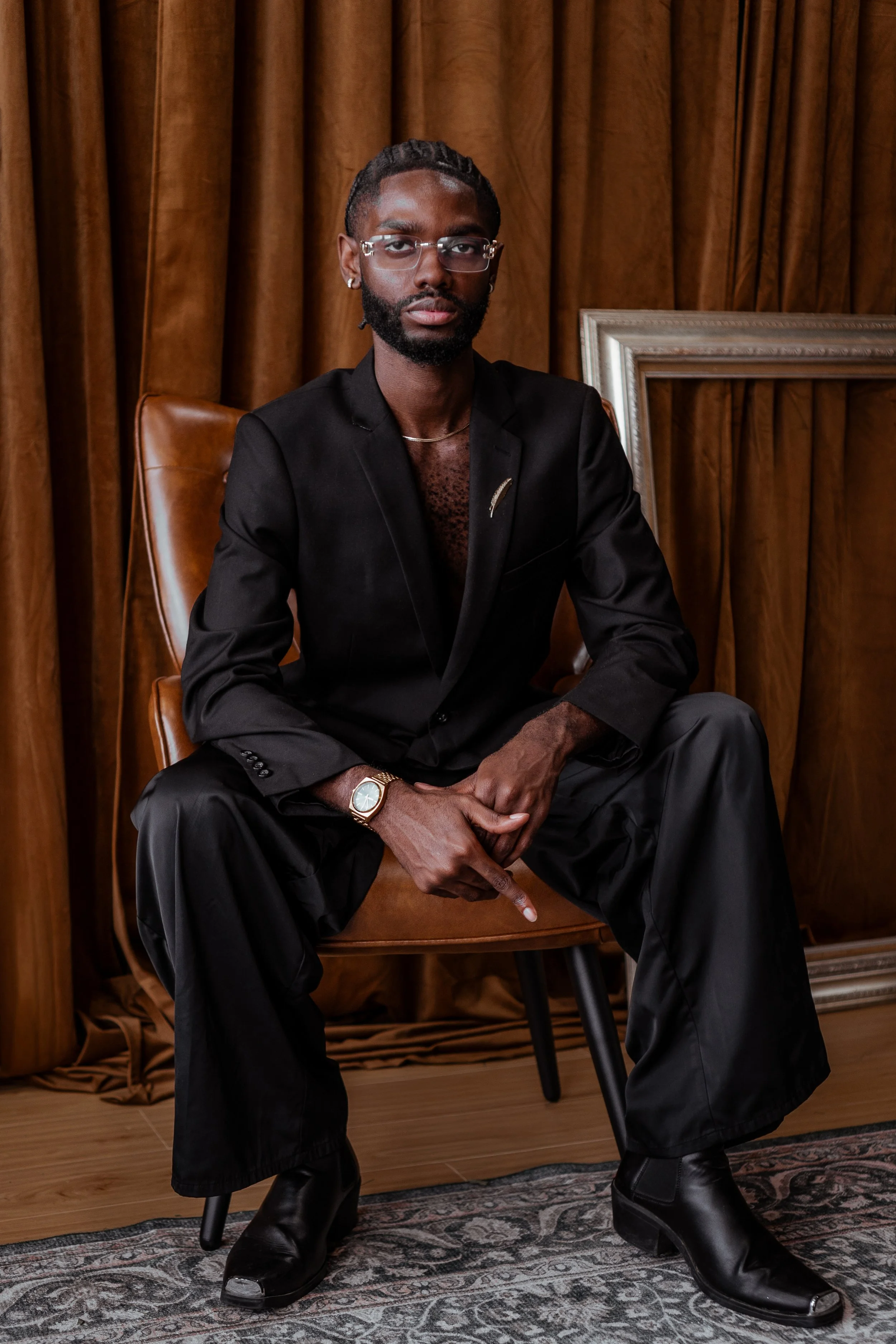 A young man with a chic black suit in a elegant room with brown velvet curtains, wood floors, and antique rug.