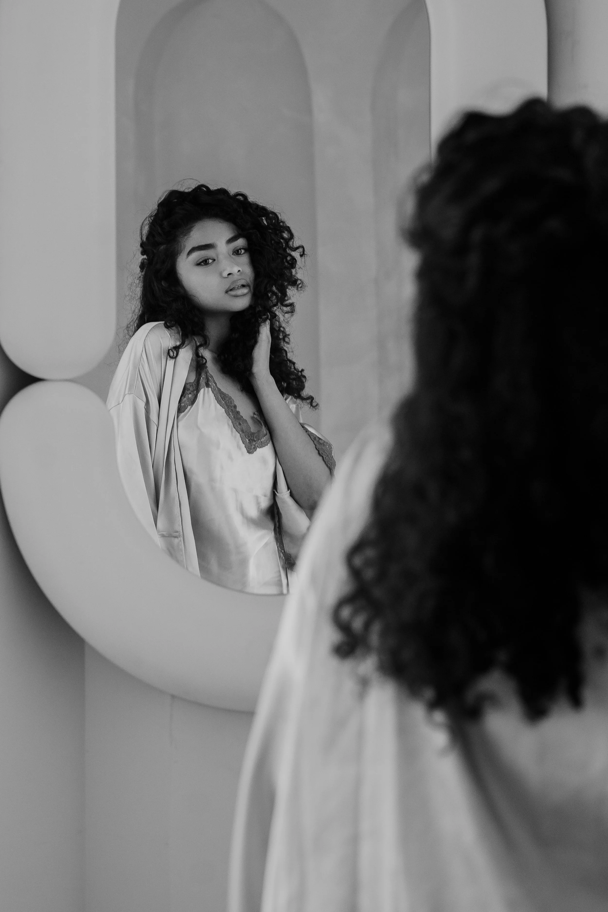 A woman with curly hair gazes into a mirror, touching her neck, wearing a satin robe with lace trim in a minimalist bathroom.