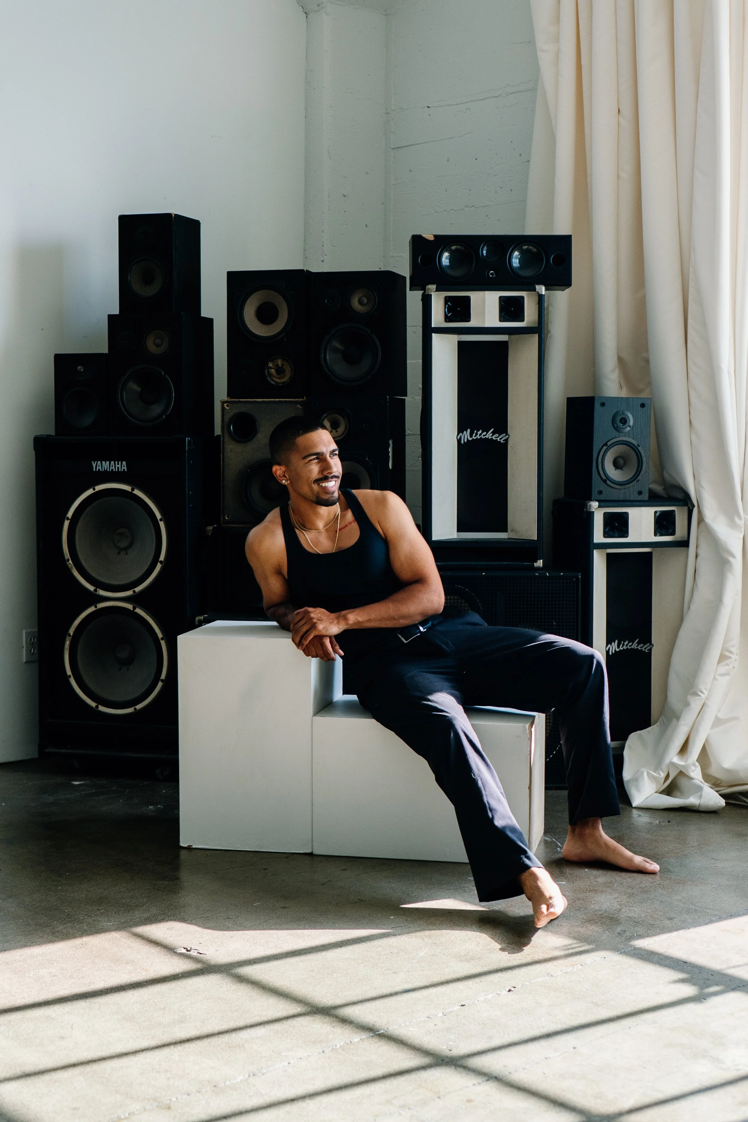 A man with short dark hair, a beard, and a smile, sitting on a white box in front of a large collection of black speakers, in a room with white walls, curtains, and sunlight casting shadows on the floor.