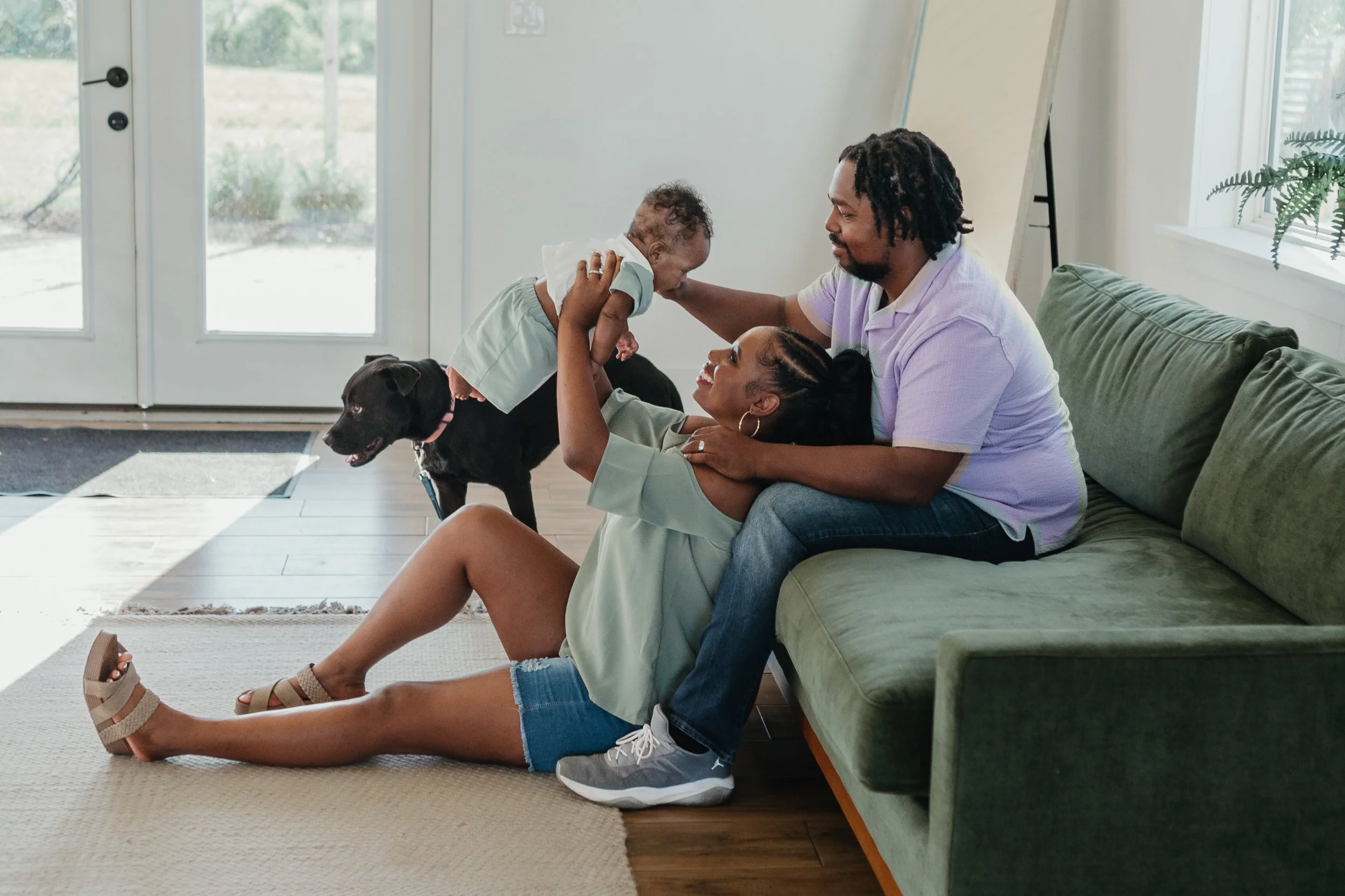 A woman and a man sitting on a green sofa inside a living room, playing with a young child who is standing by the woman. The woman is holding the child's hands while the child is leaning forward, touching the woman's nose. A dog stands beside them. L