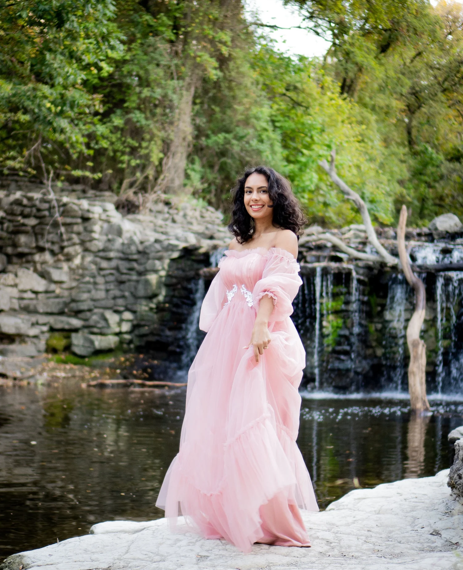 A smiling woman with curly dark hair, wearing a long pink off-shoulder dress, stands on a rocky area near a river with a forested background.