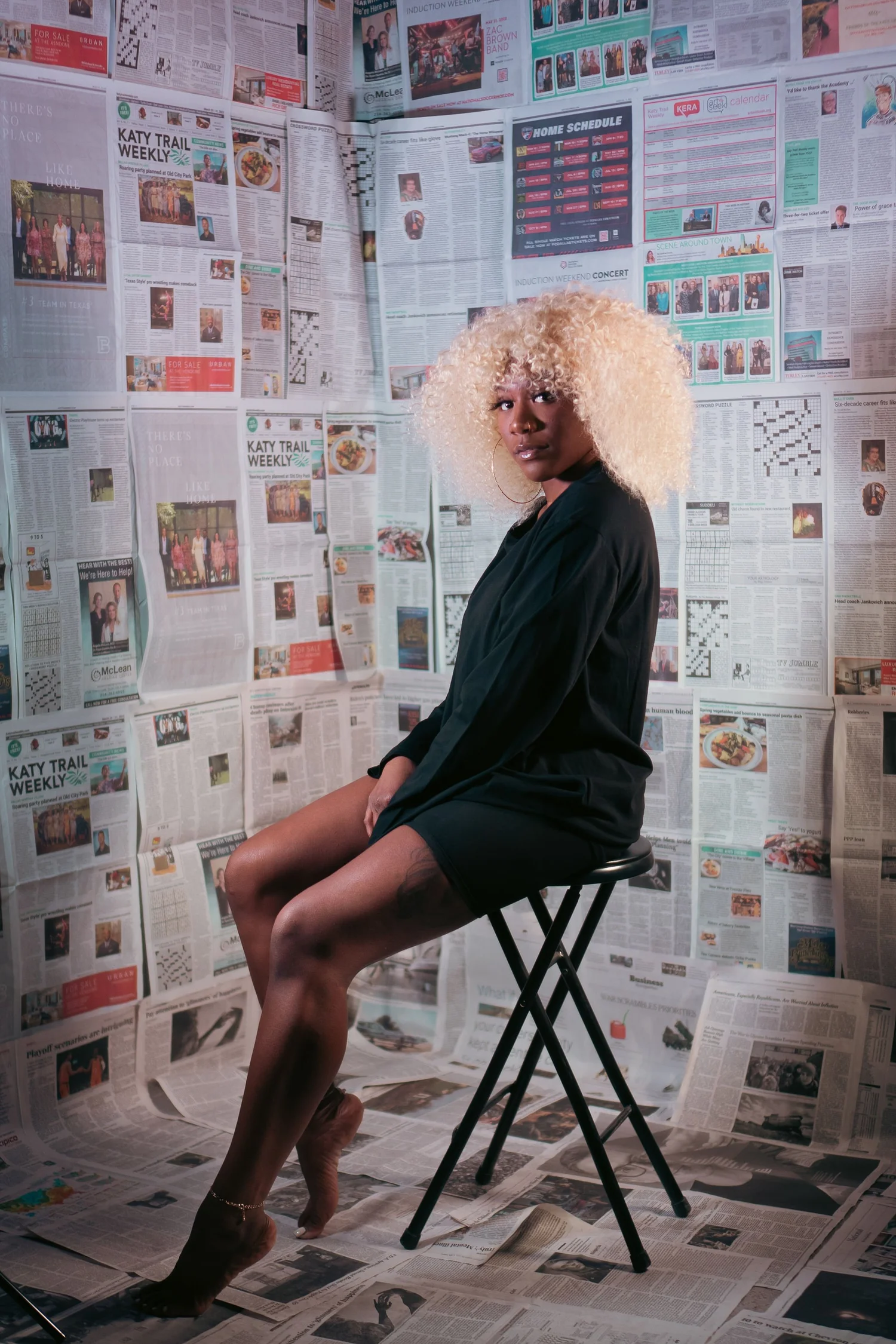 A woman with platinum blonde curly hair seated on a black stool in a room covered in newspapers on the walls and floor. She wears a black outfit and gold hoop earrings, looking directly at the camera.