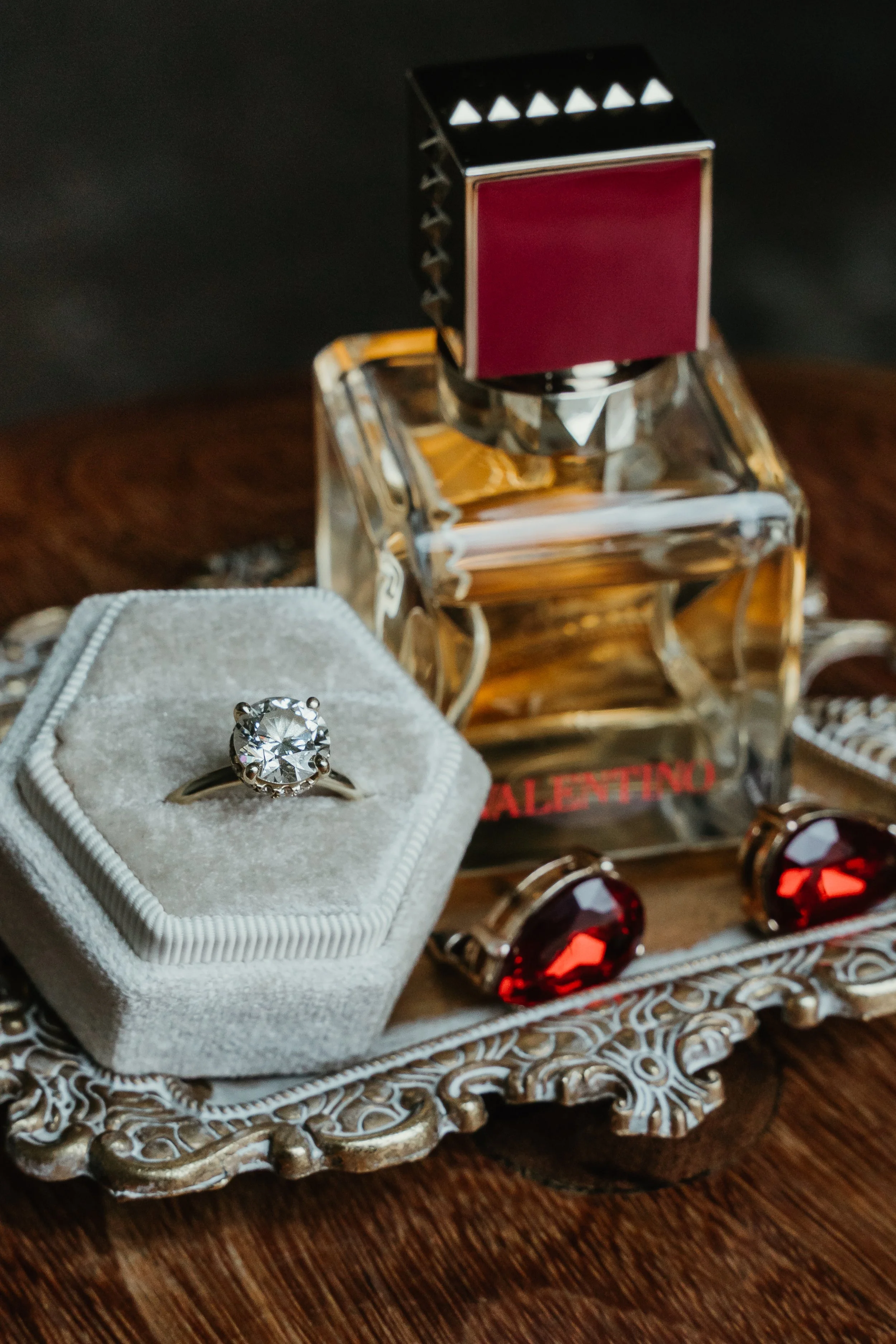 Close-up of a solitaire diamond engagement ring on a gray velvet ring box, with jewelry and a Valentino perfume bottle in the background on a decorative silver tray.