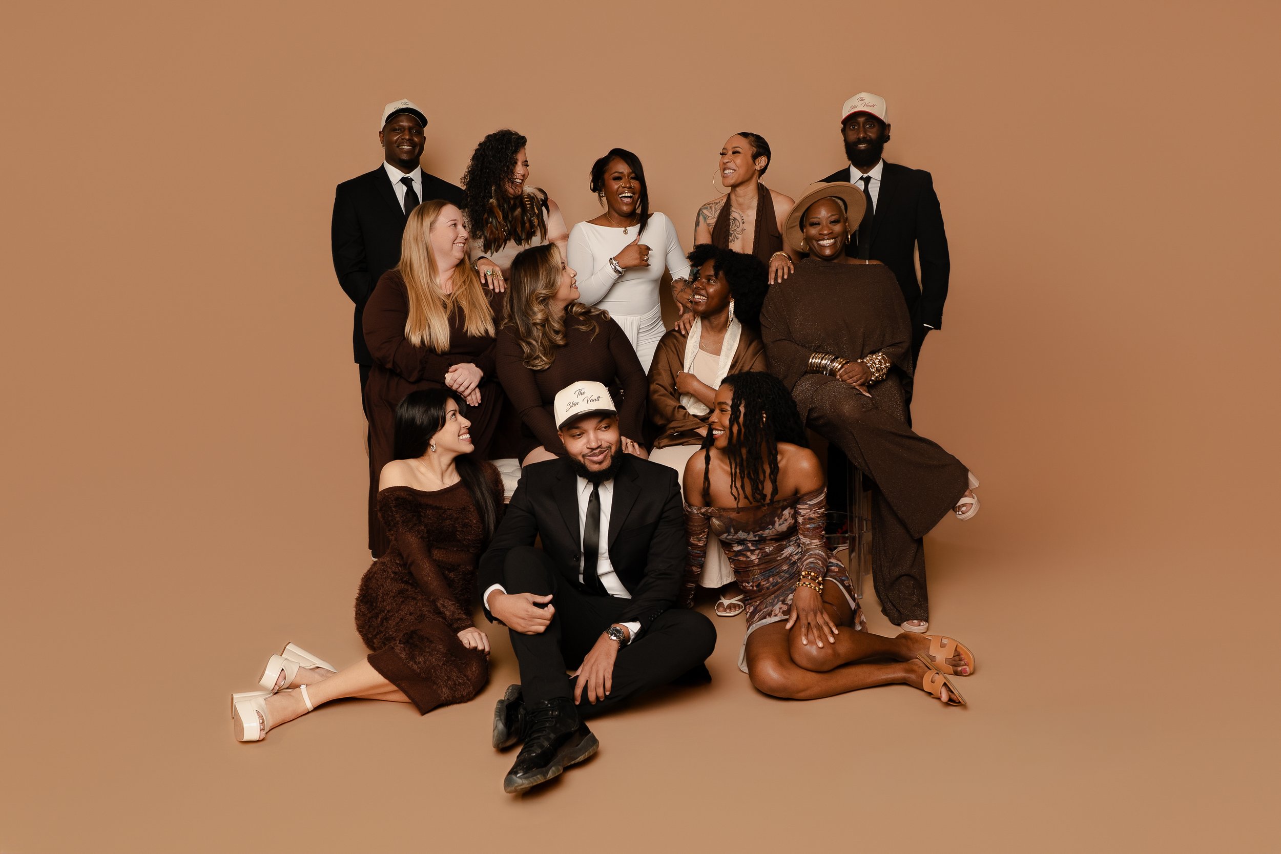 Group of diverse people laughing and smiling together against a plain beige background, some sitting and some standing, dressed in fashionable clothing.