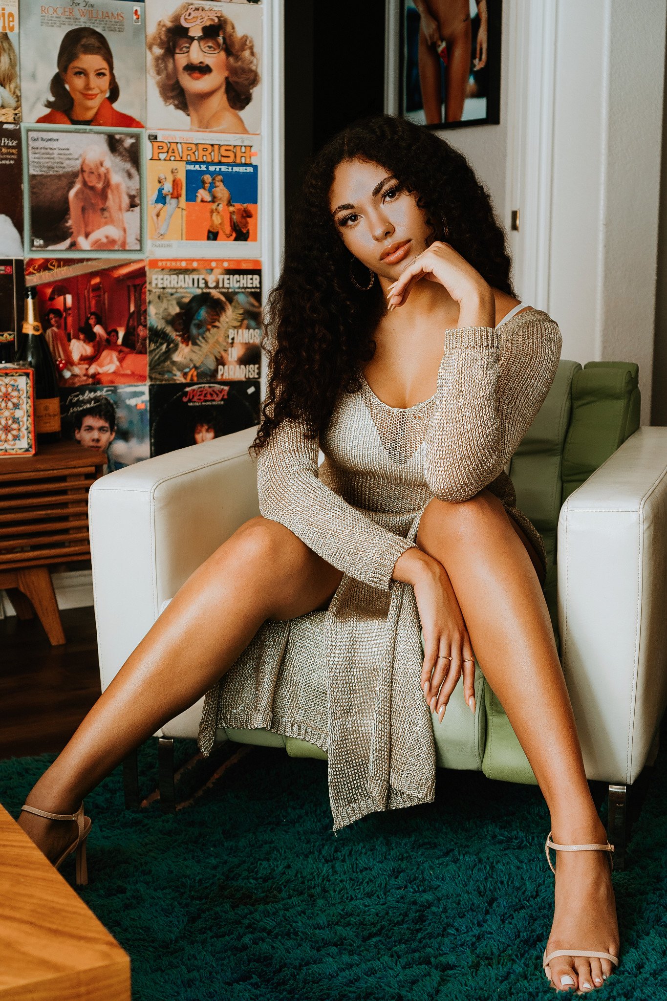 A young woman with curly dark hair and tan skin poses sitting on a couch with her legs spread apart, resting her chin on her hand, wearing a metallic gold dress and beige heels. Behind her is a wall with vintage posters and album covers, and she is s