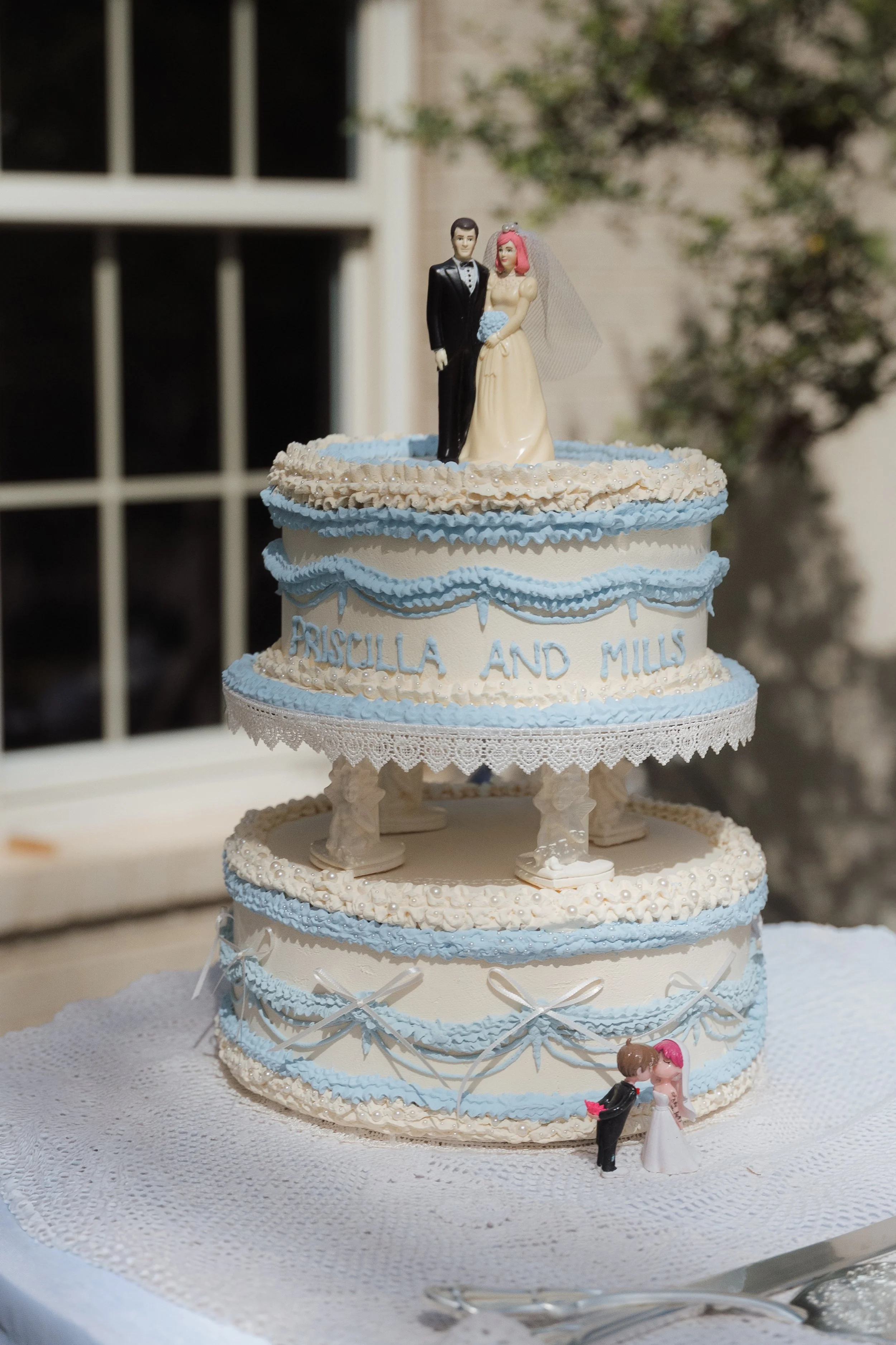A multi-layered wedding cake with blue and white decorations, topped with wedding figurines of a bride and groom, and decorated with small floral and bow details.