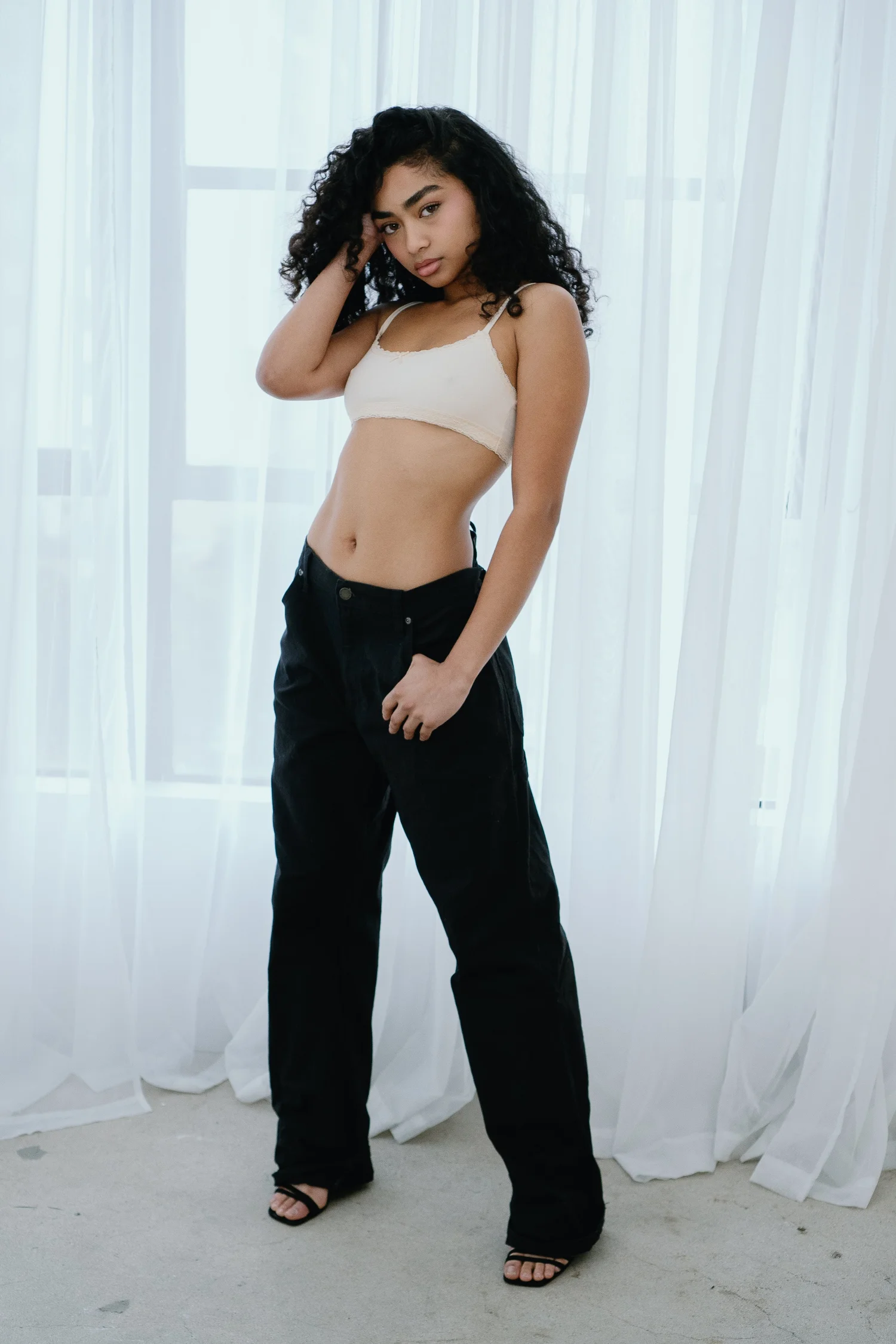 Young woman with curly hair in a white crop top and black pants standing indoors near curtains.