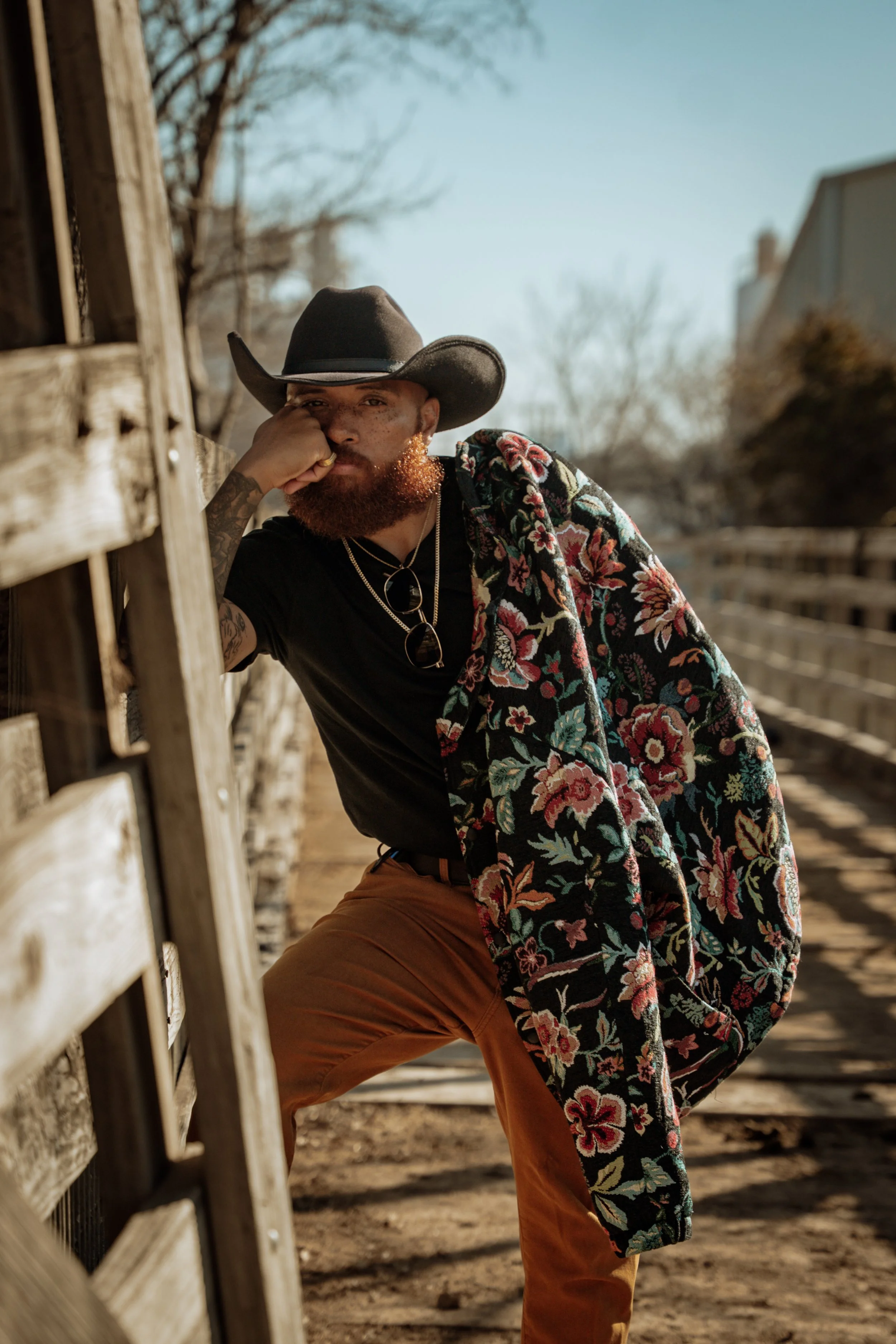 A man with a red beard and tattoos wearing a cowboy hat, black t-shirt, and a floral jacket leaning on a wooden fence outdoors on a sunny day.