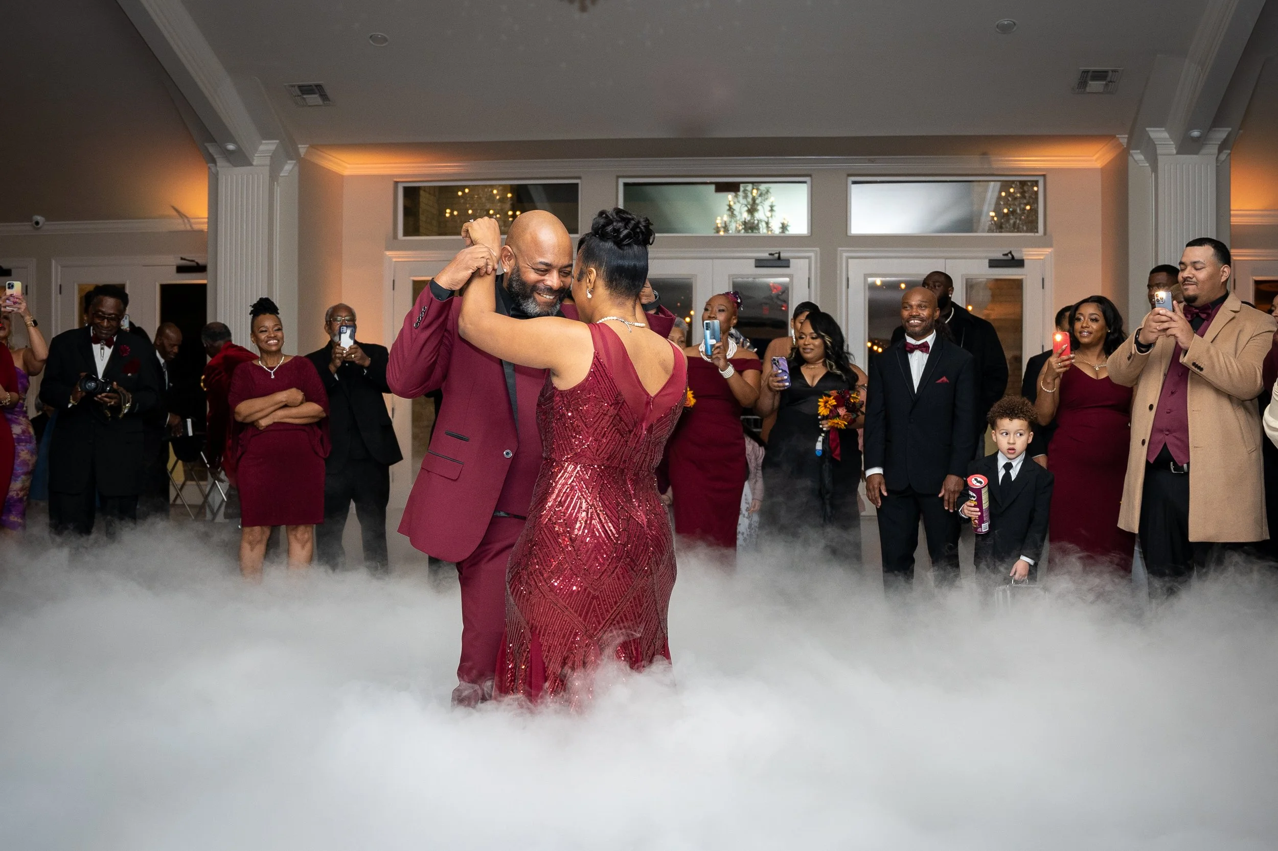 A couple dances closely at a formal event while surrounded by guests taking photos and videos. The woman wears a red gown and the man a matching red suit. The group is indoors, with festive decorations and a cloud-like fog on the floor.