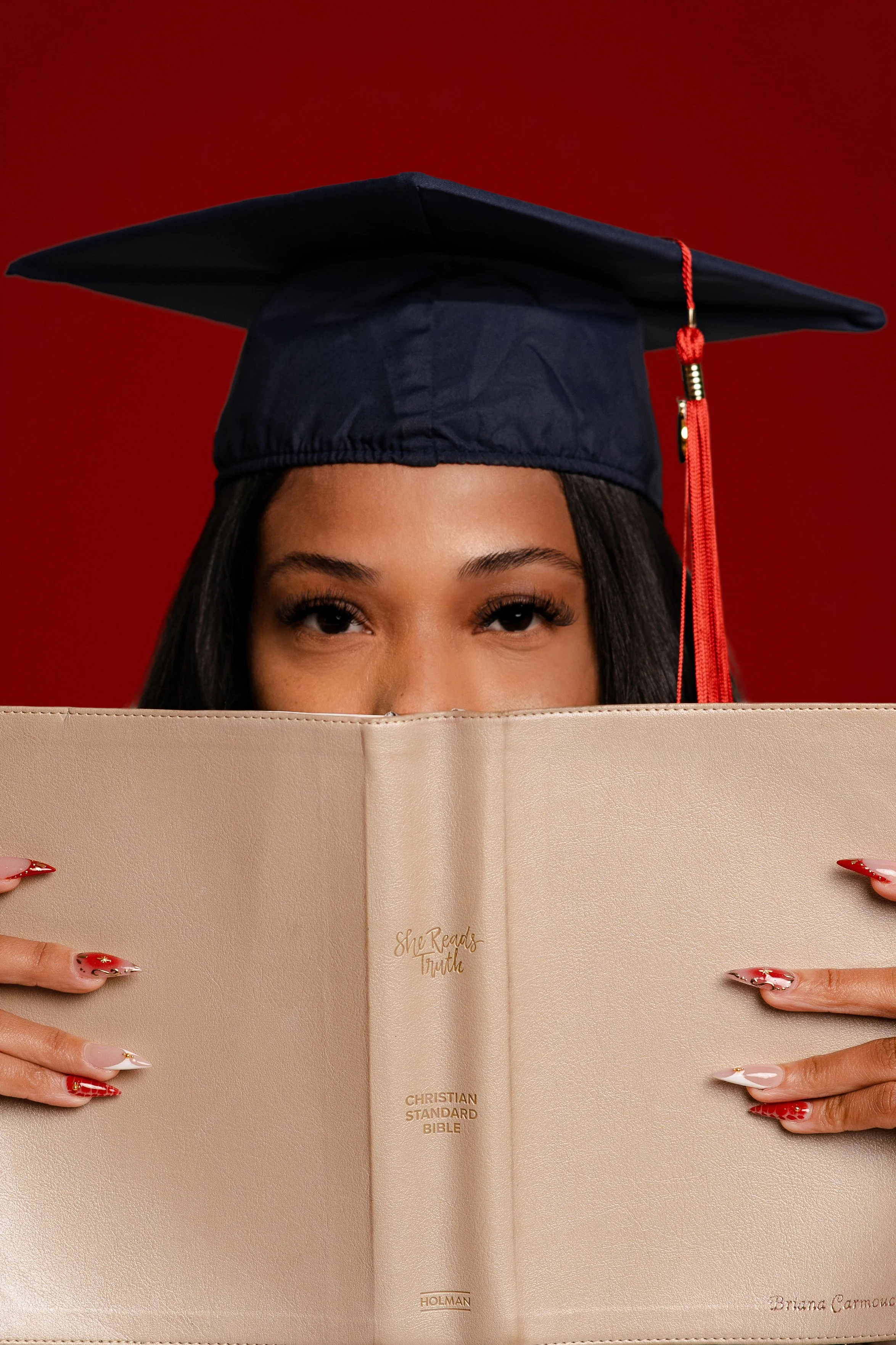 A woman with long dark hair and manicured nails holds a beige Christian Standard Bible in front of her, with only her eyes and forehead visible, wearing a dark graduation cap with red tassel, against a red background.