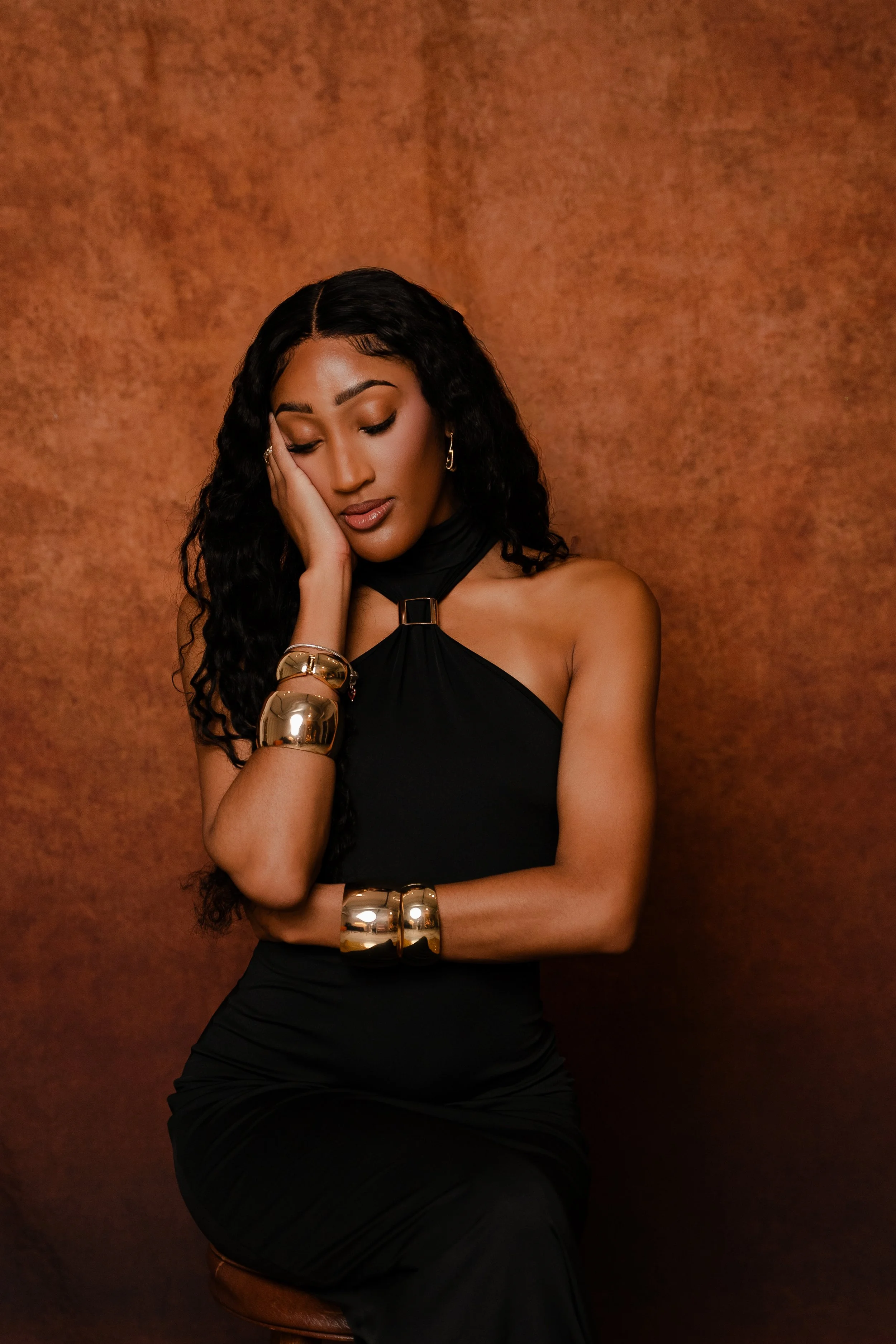 A woman with long, curly black hair is wearing a black sleeveless dress, gold bracelets, earrings, and is resting her head on her hand against a brown textured background.