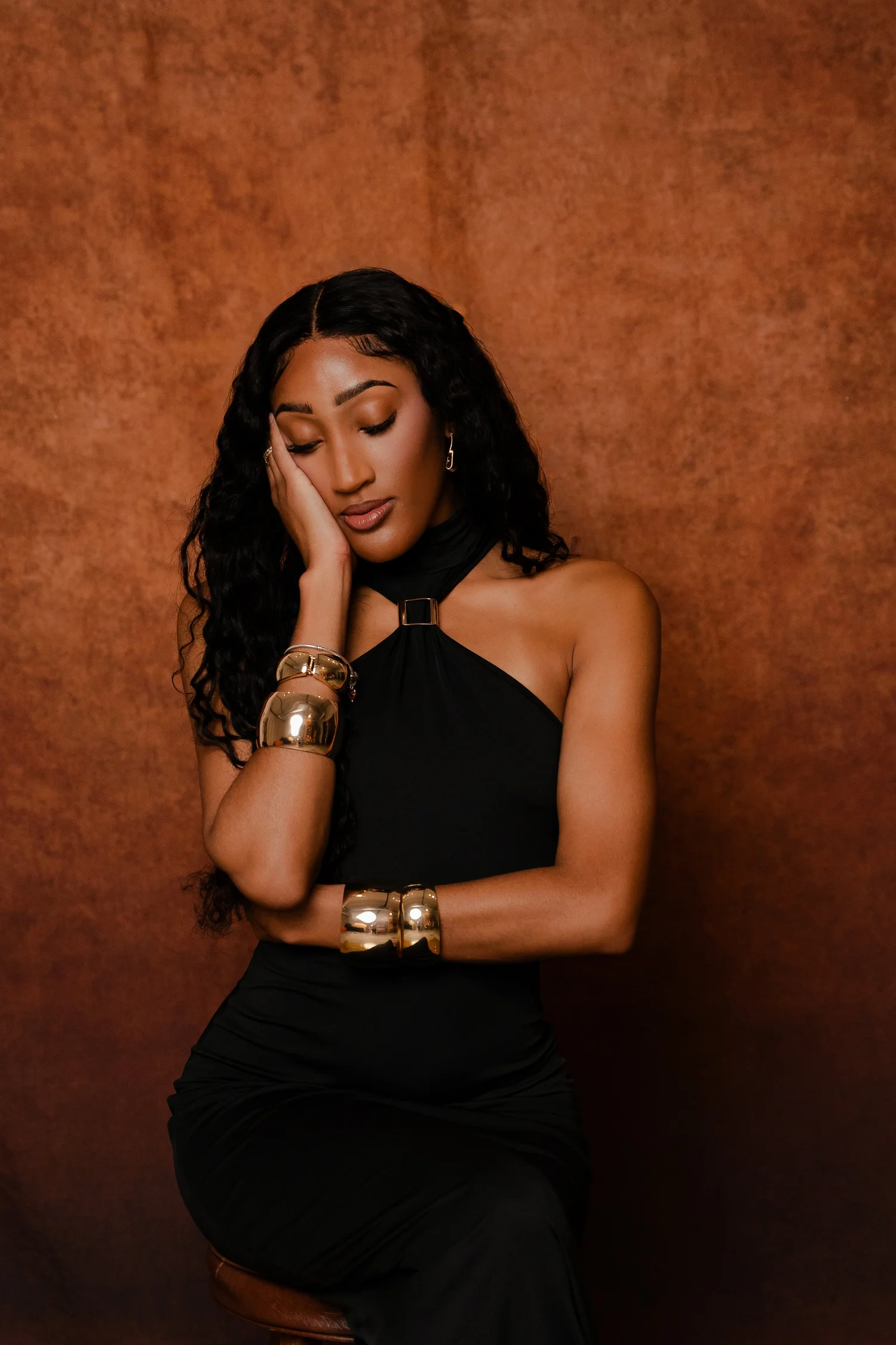A woman with long, curly black hair is wearing a black sleeveless dress, gold bracelets, earrings, and is resting her head on her hand against a brown textured background in a studio in Arlington, TX.