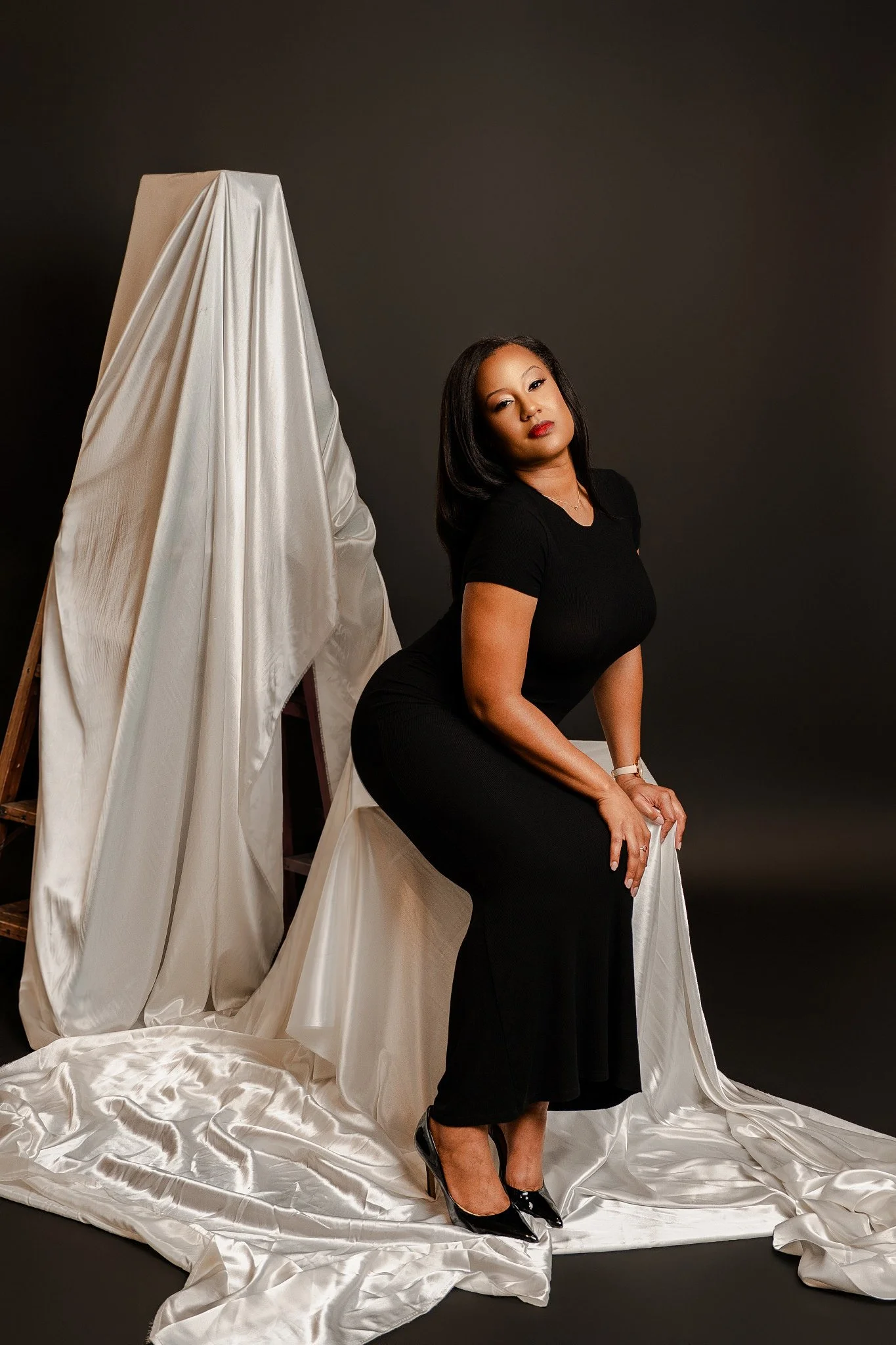A woman in a black dress and high heels sits on a white fabric-covered chair, posing with a serious expression against a dark background, with draped white satin fabric on the floor and surrounding her.