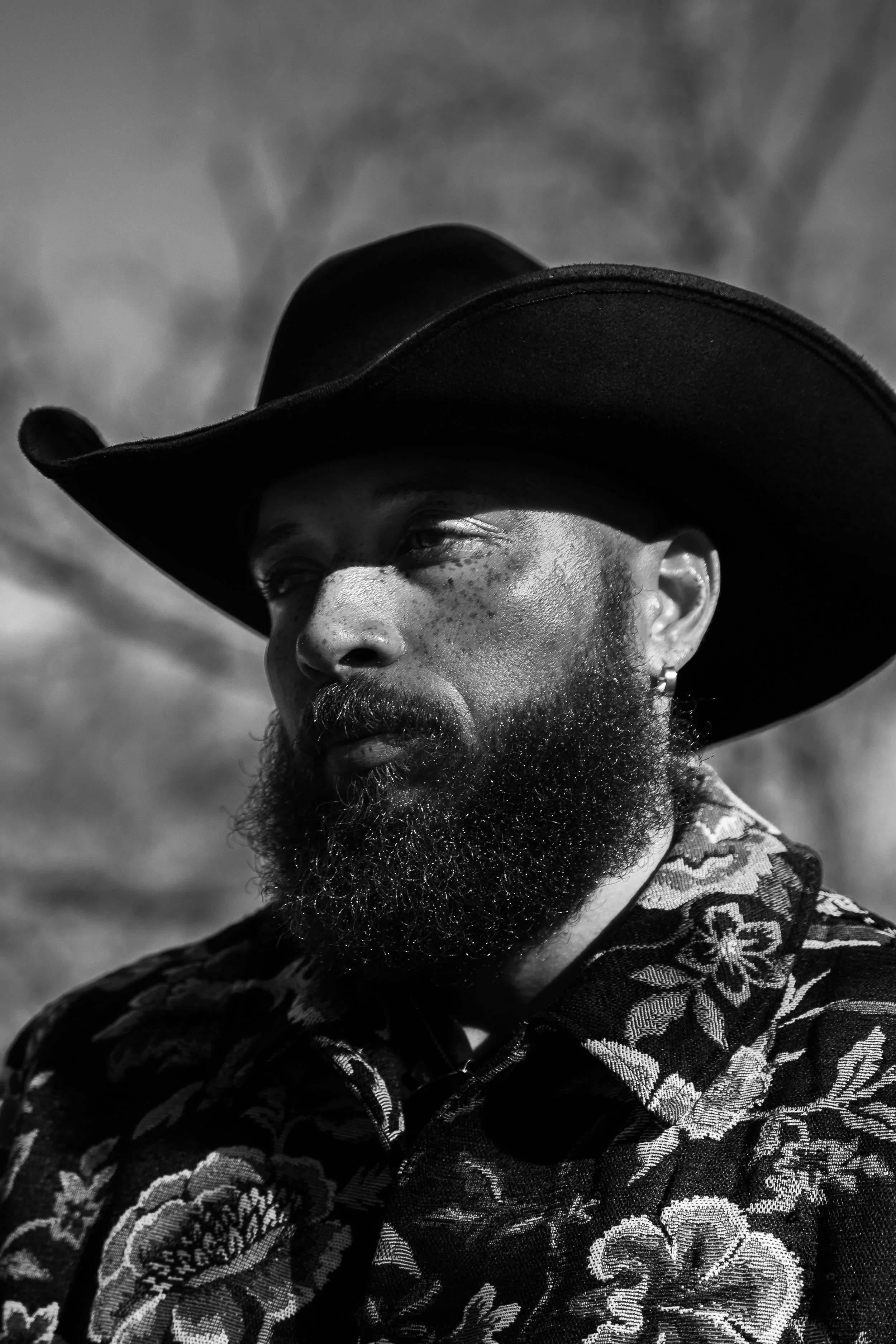 Black and white photo of a man with a beard wearing a large wide-brimmed hat and patterned jacket, with clouds in the background.