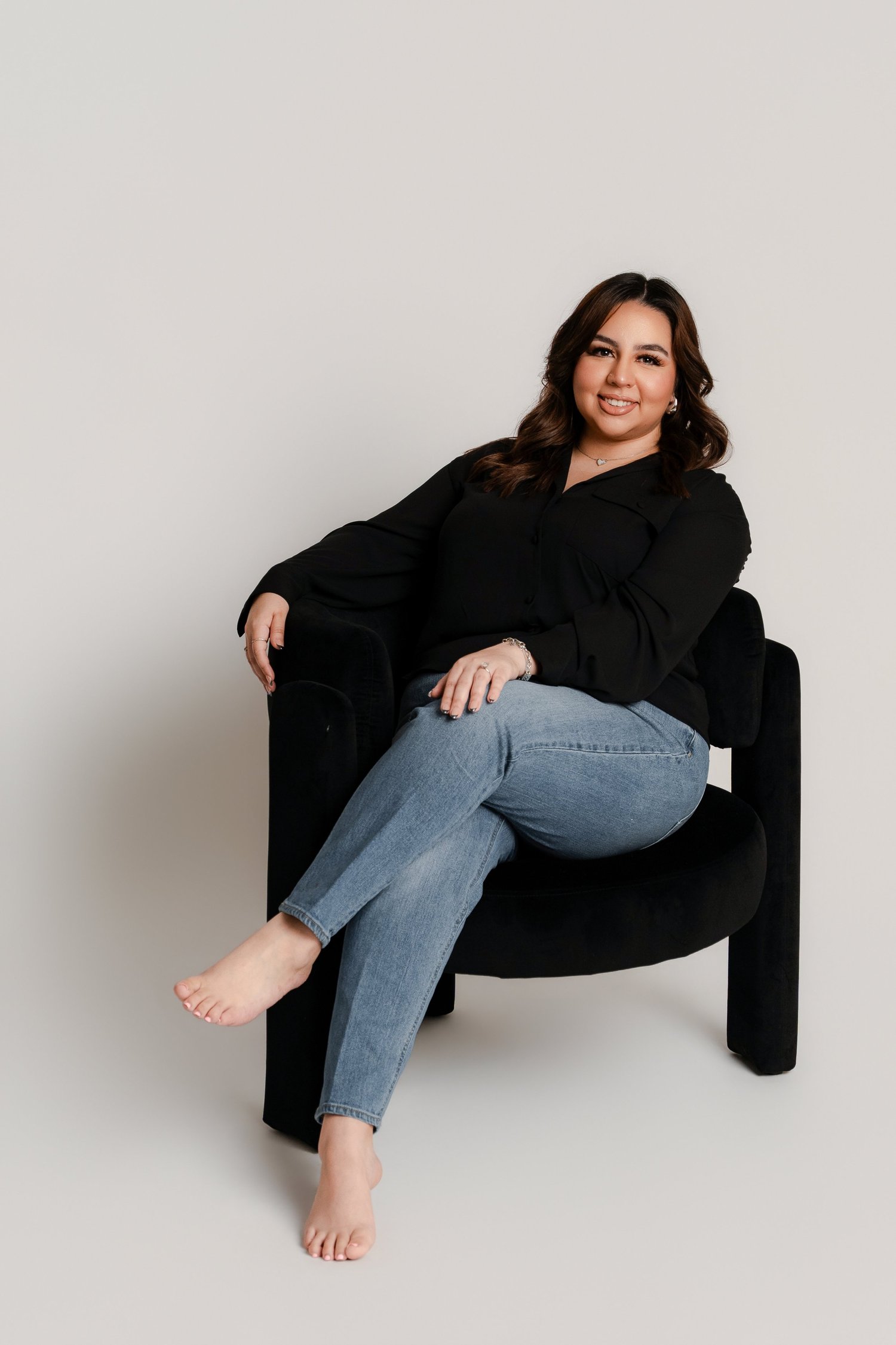 A woman with brown hair, wearing a black top and light blue jeans, sitting barefoot on a black chair, smiling in front of a plain white wall.