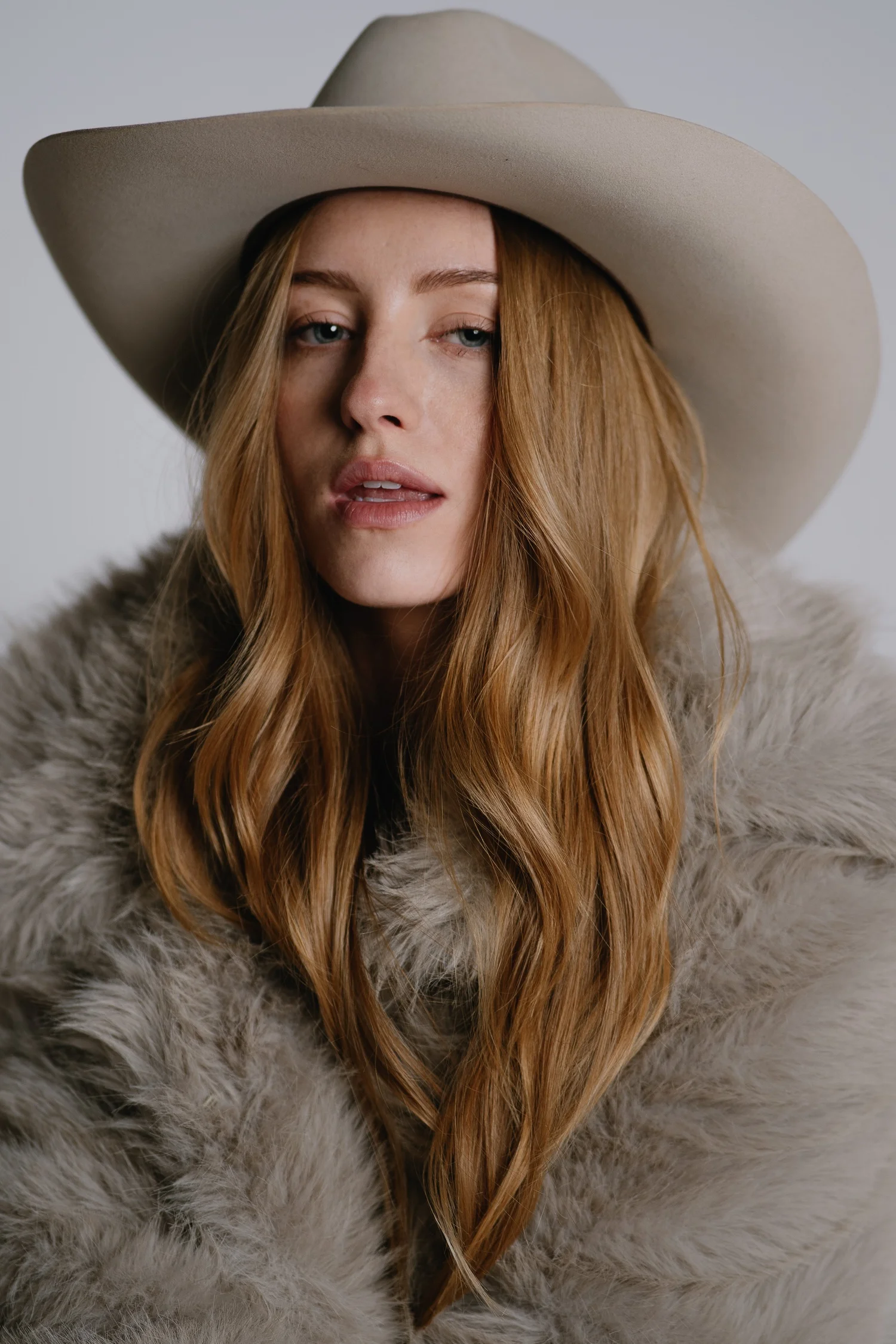 A woman with long red hair wearing a beige wide-brimmed hat and a fur coat.