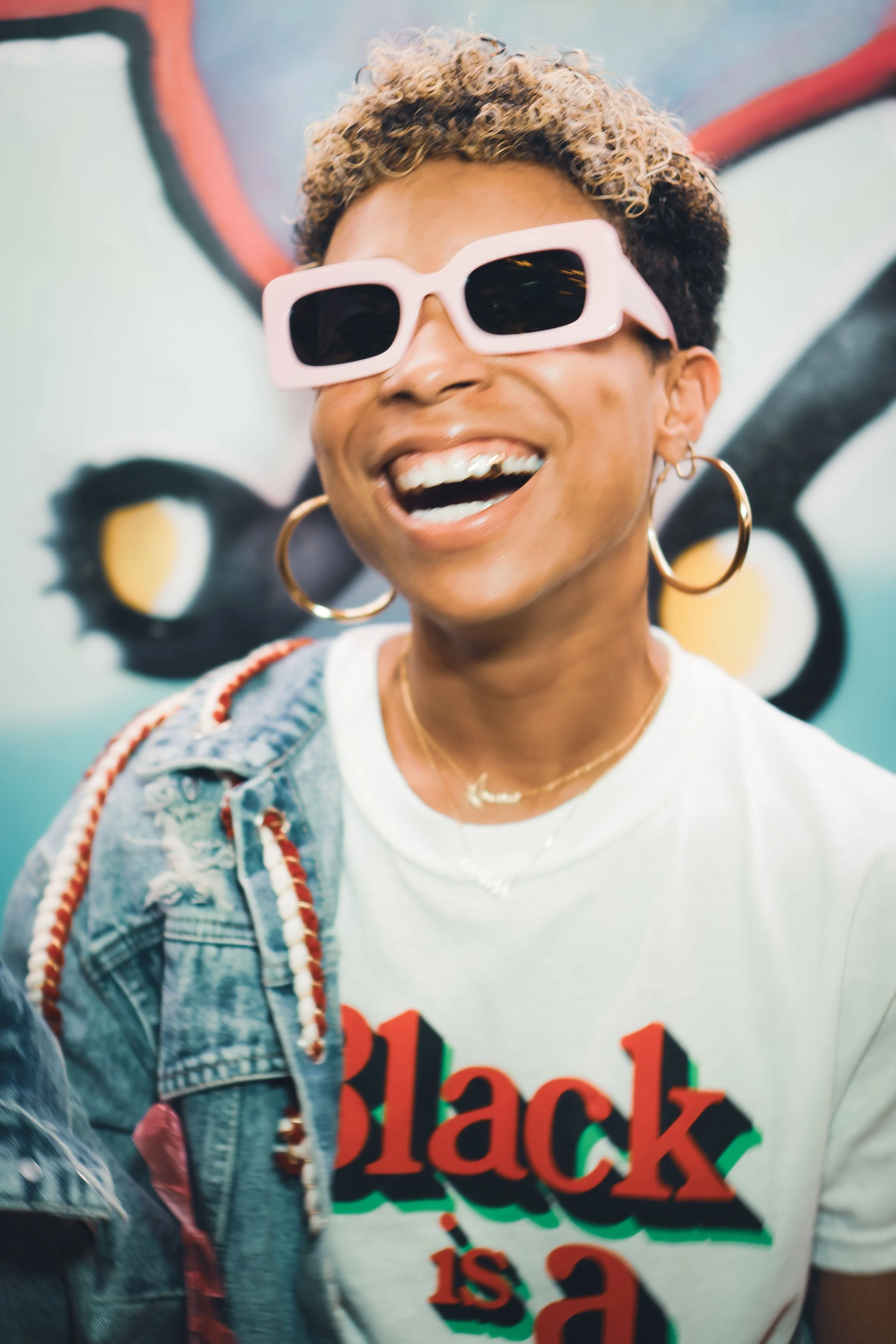 A person with curly hair wearing large pink sunglasses, gold hoop earrings, and a white t-shirt with a colorful graphic reading 'Black is a', partially visible, standing in front of a graffiti wall.