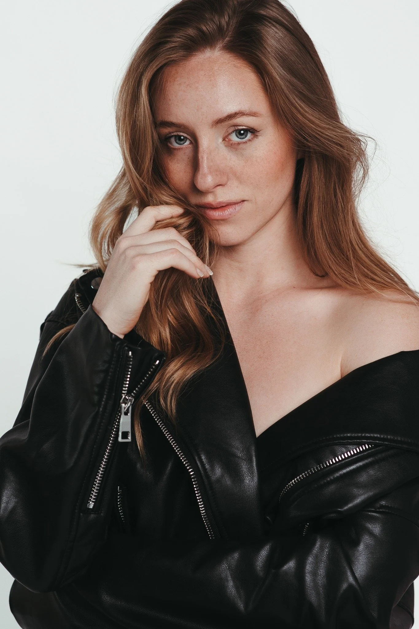 Editorial studio portrait of a red-haired woman in a black leather jacket against a pale background.