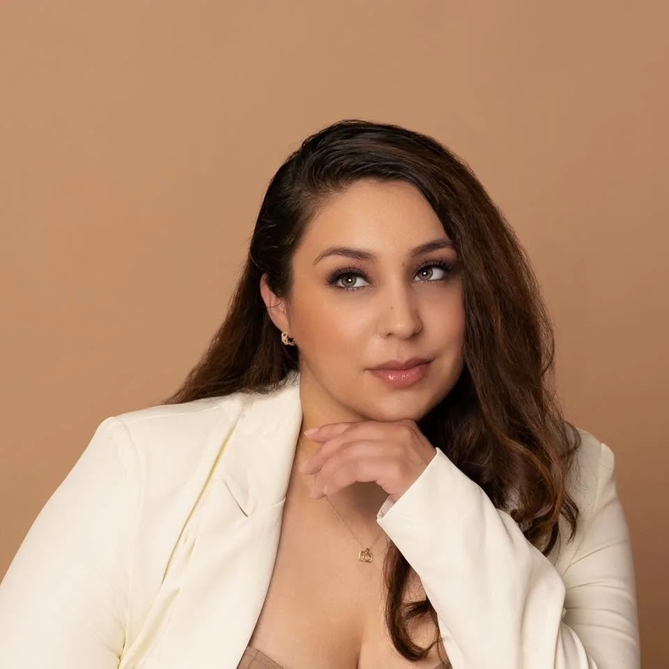 A woman with long brown hair and light makeup, wearing a white blazer and gold jewelry, posing against a light brown background.