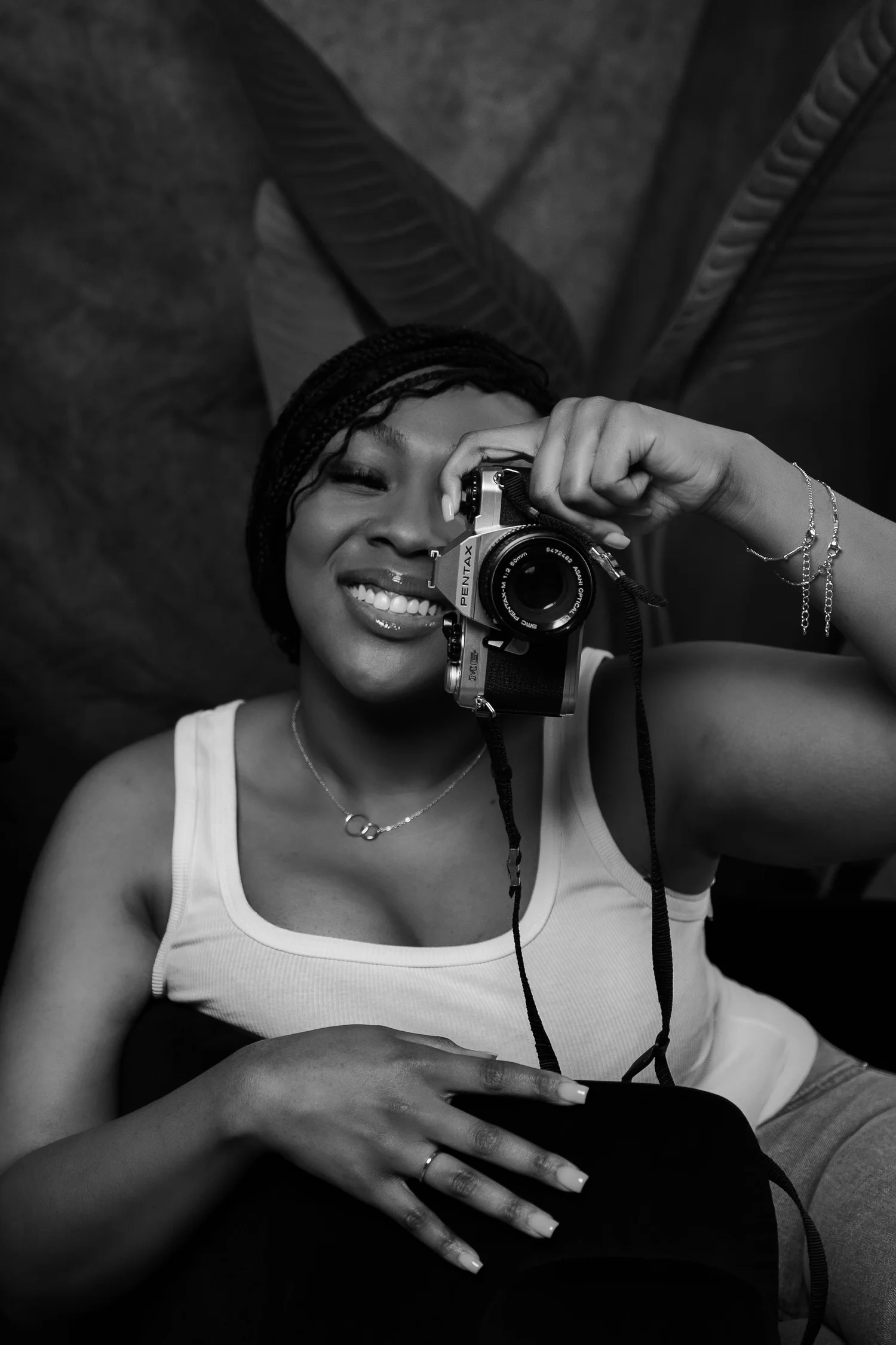 A woman with braided hair smiling while taking a selfie with a camera in a mirror. She is wearing a sleeveless top, a necklace, and several bracelets. The background features large plant leaves.