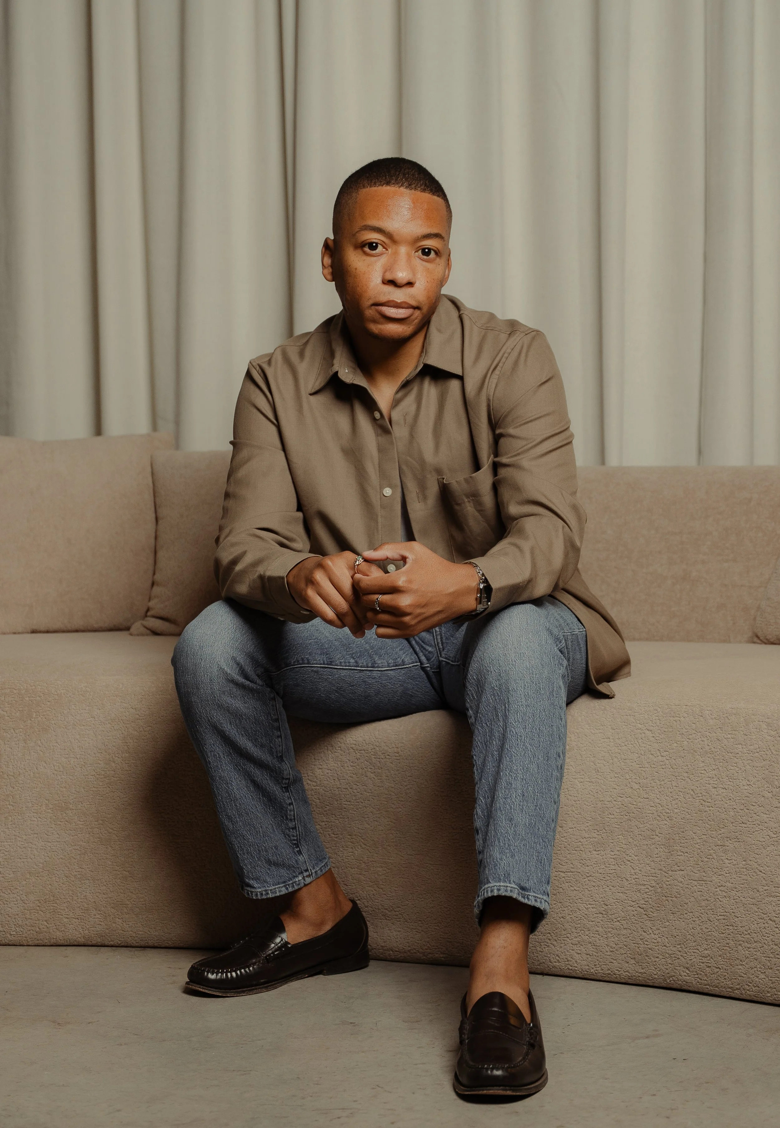 A person sitting on a beige couch, wearing a brown button-up shirt, blue jeans, and black loafers, with blond hair and looking at the camera in front of beige curtains.