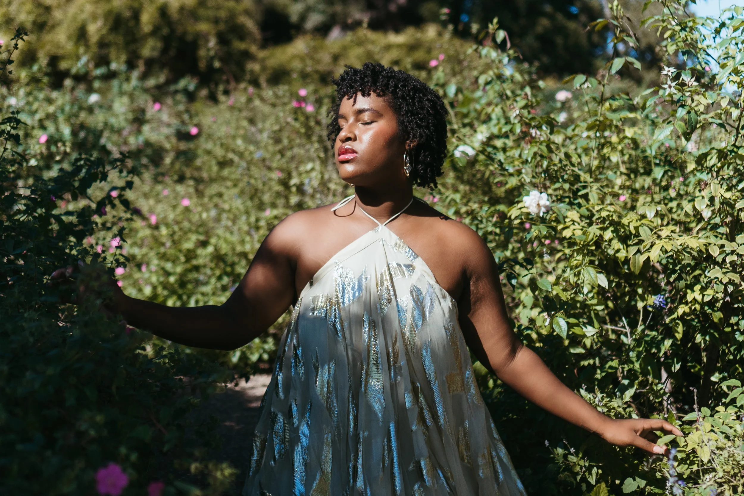 A woman with dark curly hair, closed eyes, and a calm expression, standing outdoors among greenery and flowering bushes, wearing a sleeveless white and silver dress, with sunlight highlighting her face and shoulders.