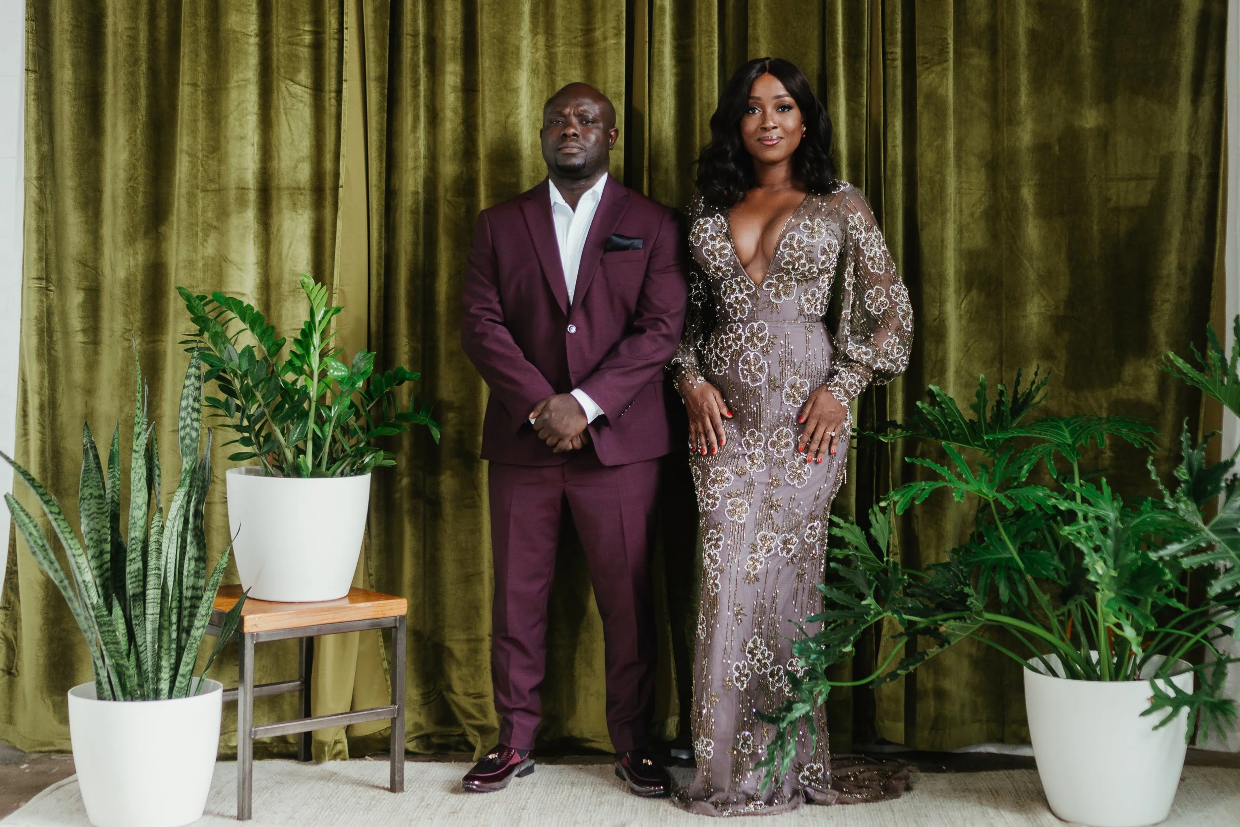 A man in a maroon suit and a woman in a floral evening gown standing in front of a green curtain, surrounded by potted plants.