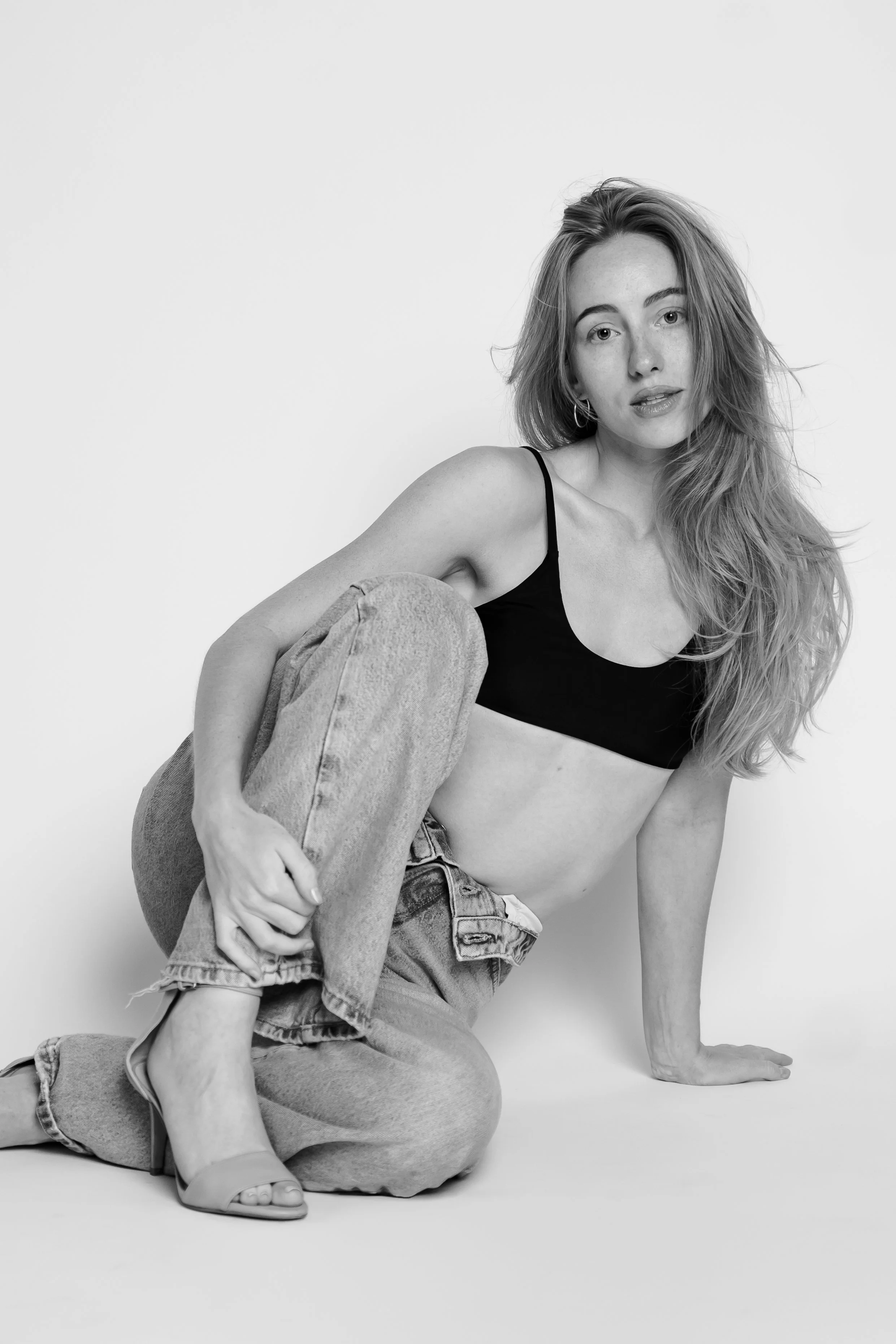 Black and white photo of a young woman with long hair, wearing a black crop top, jeans, and high heels, crouching on the floor against a plain background.