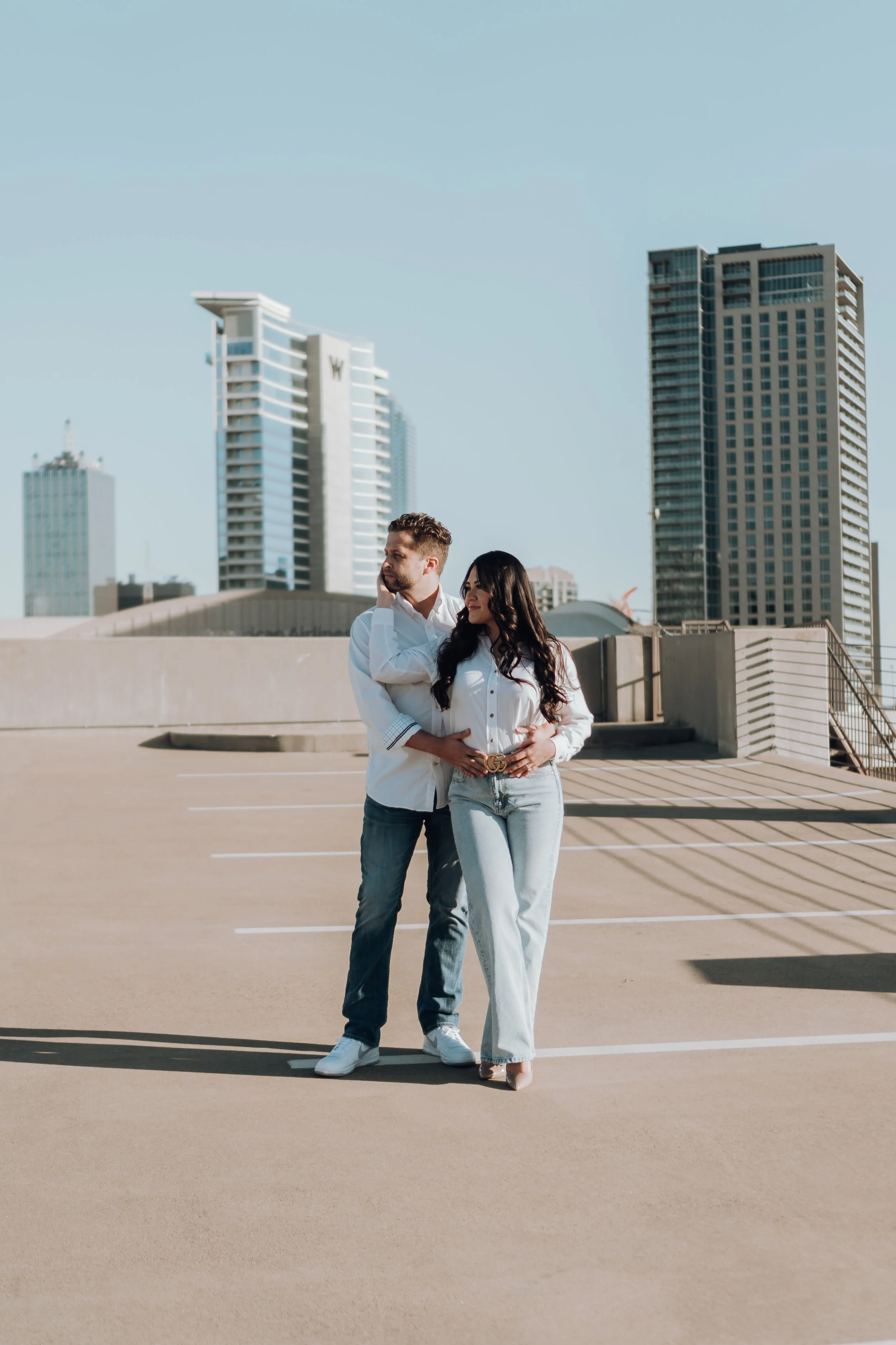 A man and woman standing close together in an empty rooftop parking lot with tall city buildings in the background. The man is dressed in a white shirt and jeans, and the woman is in a white blouse and light blue jeans.