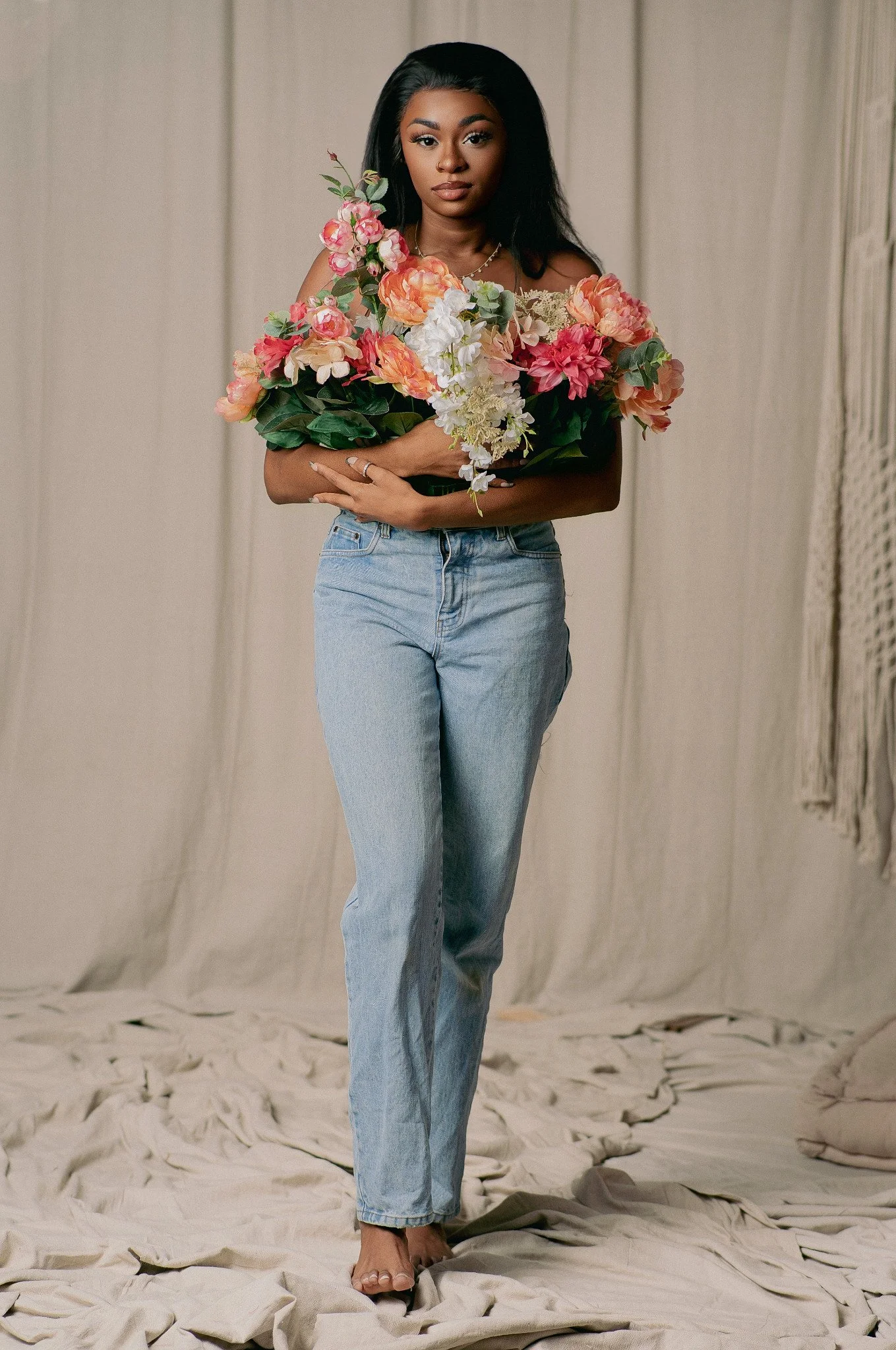A woman with dark hair and dark skin holding a large bouquet of colorful flowers, standing on a beige fabric backdrop with a textured wall on the right side.