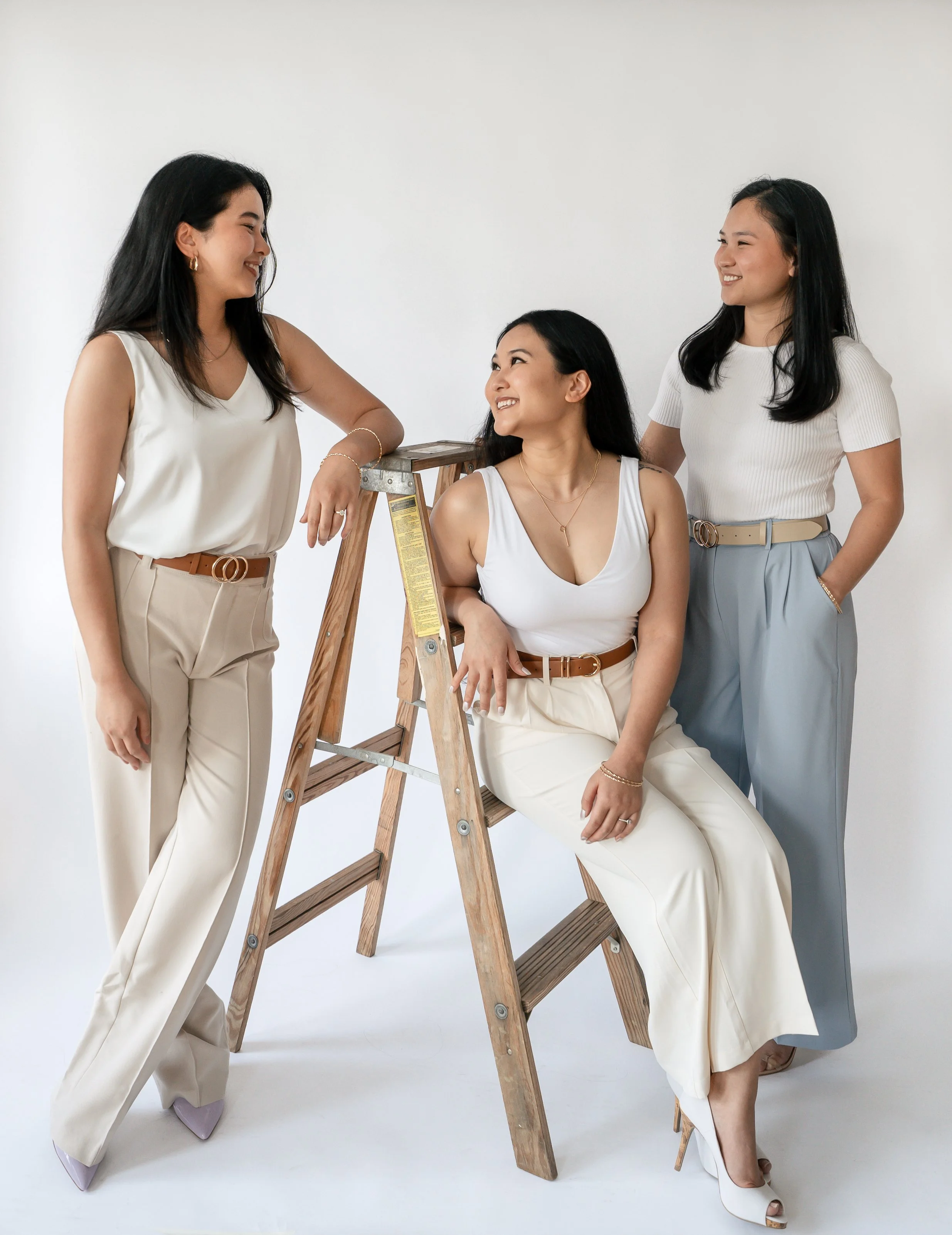 Four women dressed in neutral tones interacting around a wooden ladder, with one woman sitting on the bottom step, two women standing and leaning on the ladder, and one woman standing beside it, all smiling at each other in a studio with a white back