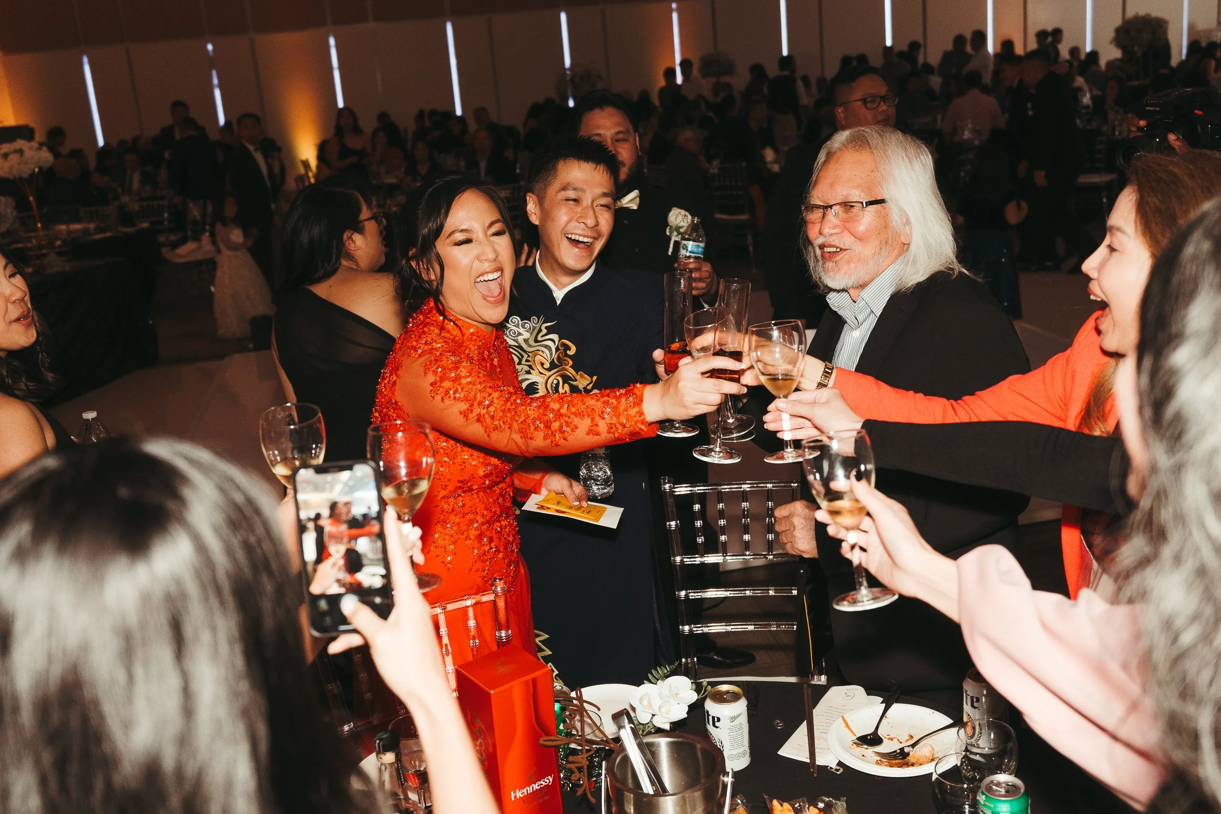 A group of people at a formal celebration or dinner party, with some raising glasses for a toast, in a large banquet hall with many guests in the background.