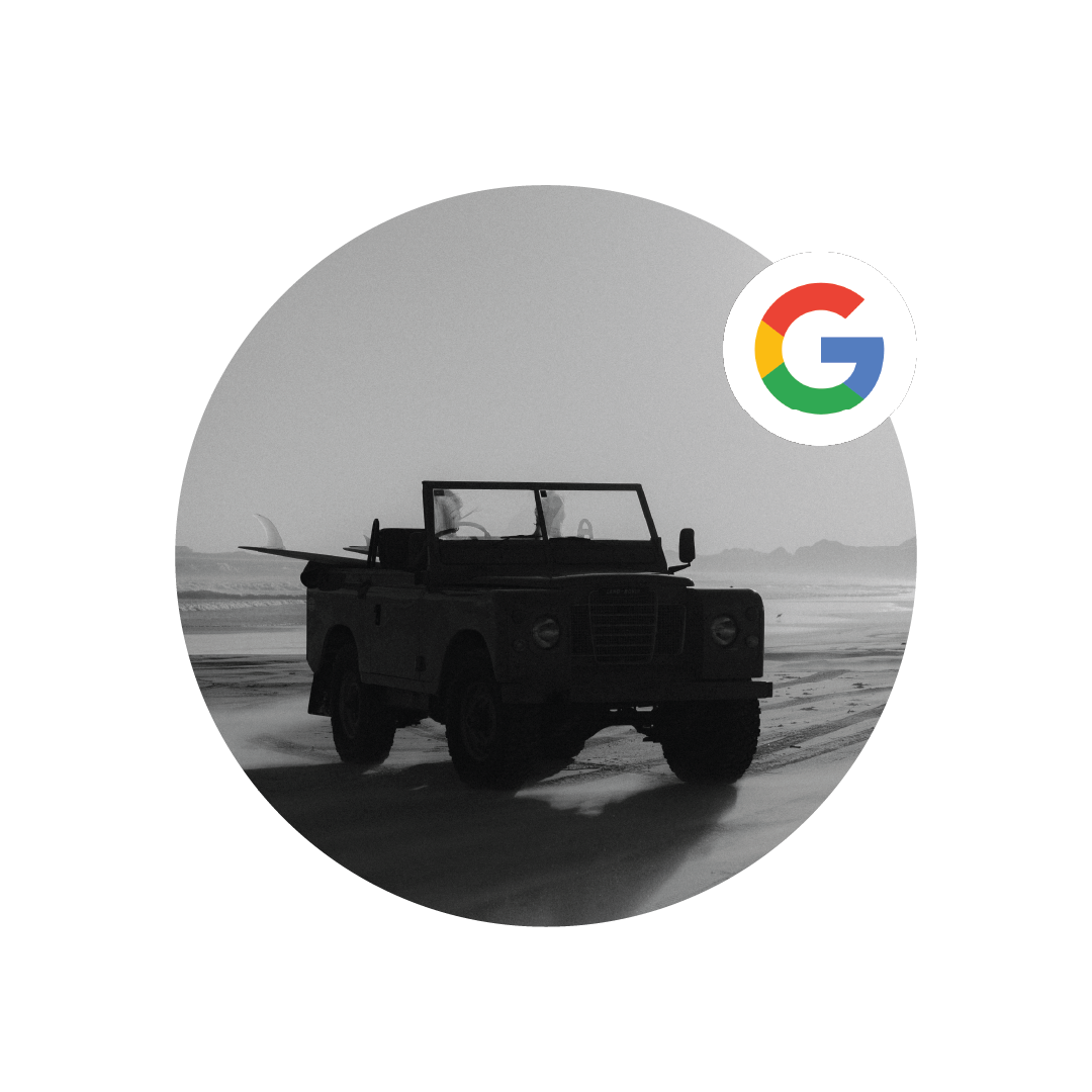 A black vintage off-road vehicle with a surfboard on its back parked on a beach with ocean waves and a mountain range in the background, in grayscale. Google logo overlay.